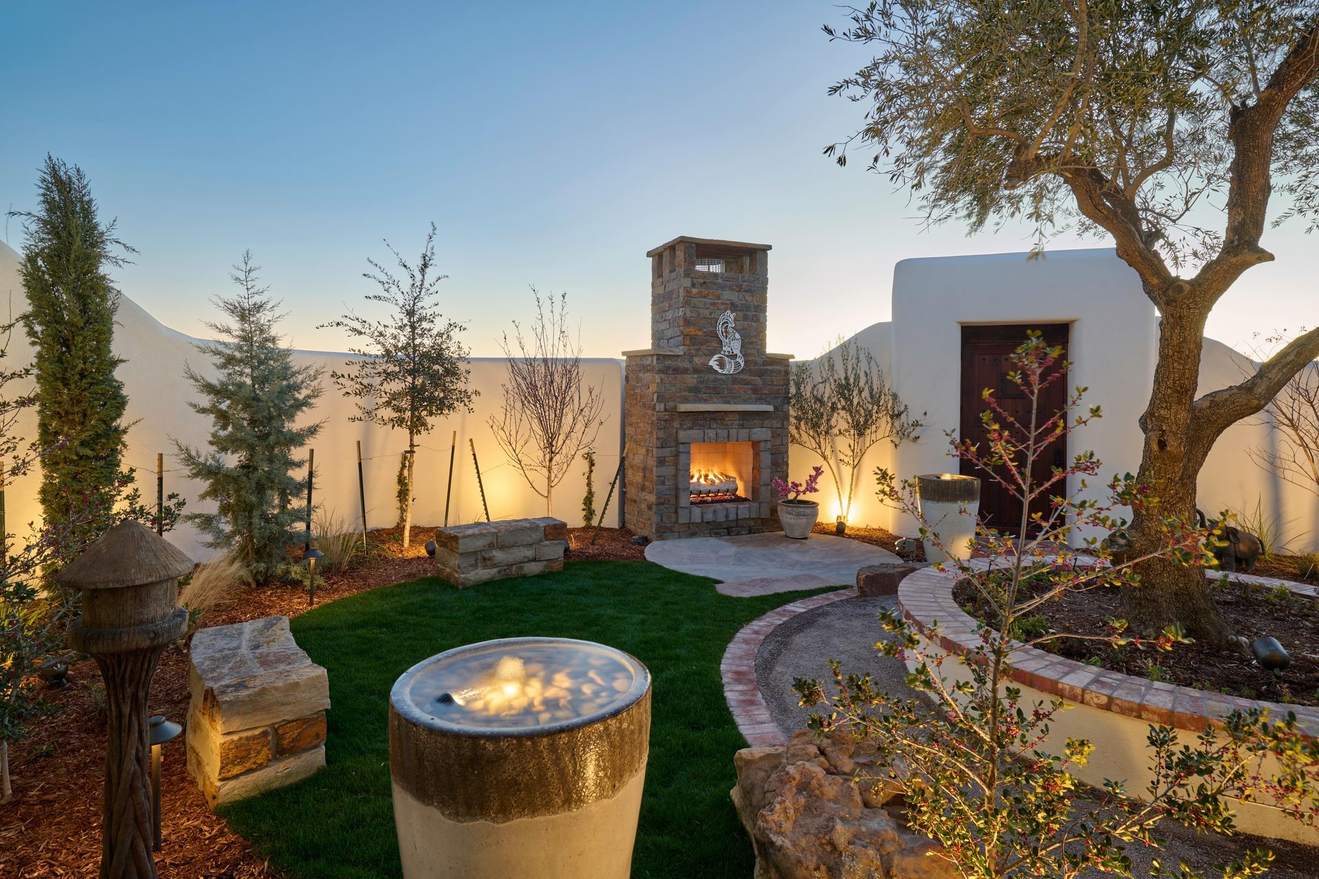 Outdoor backyard with fireplace, seating, and illuminated landscaping at dusk.