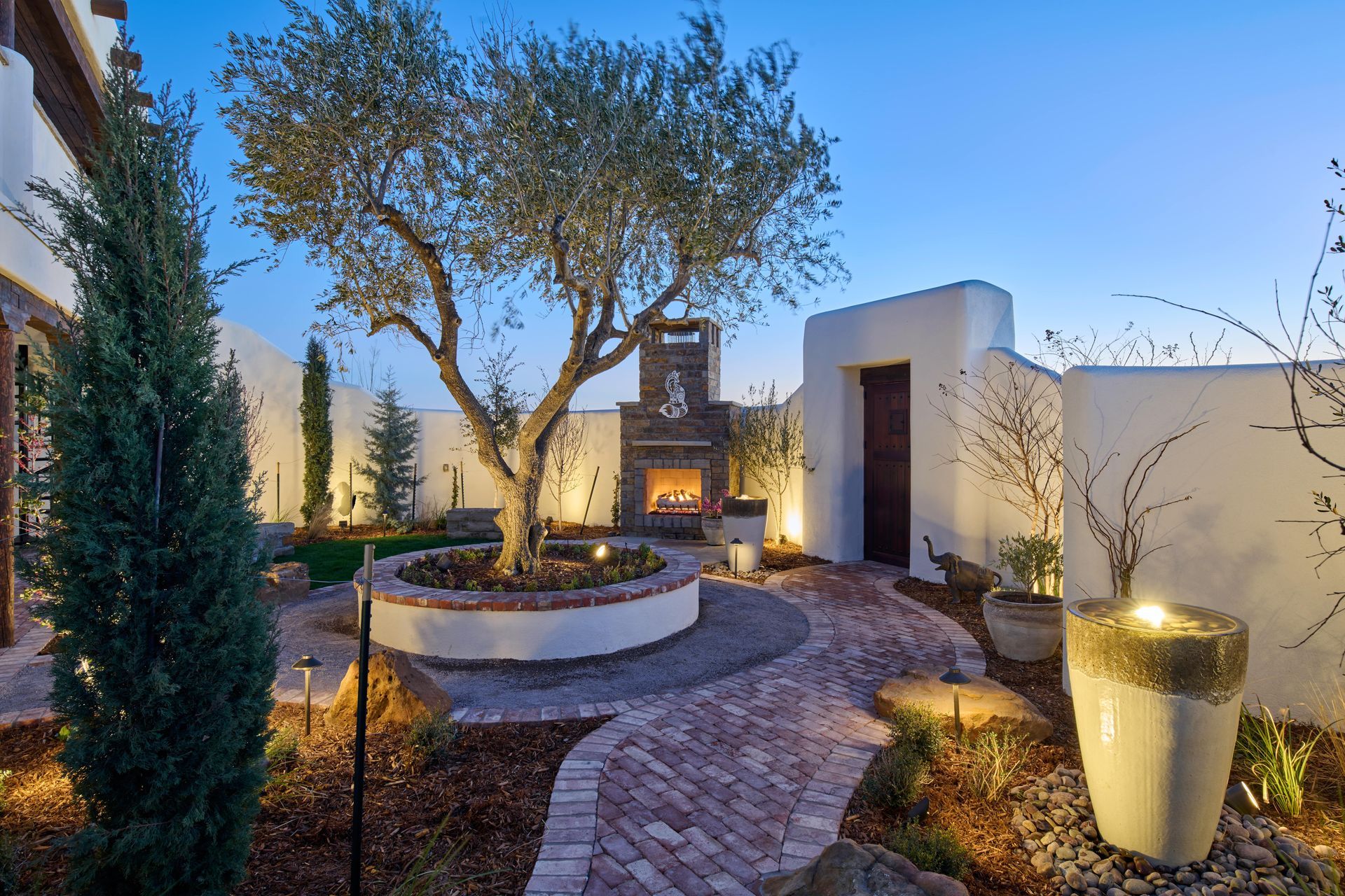 Brick pathway leads to an outdoor fireplace, with a large tree and water fountain in a walled courtyard, at dusk.