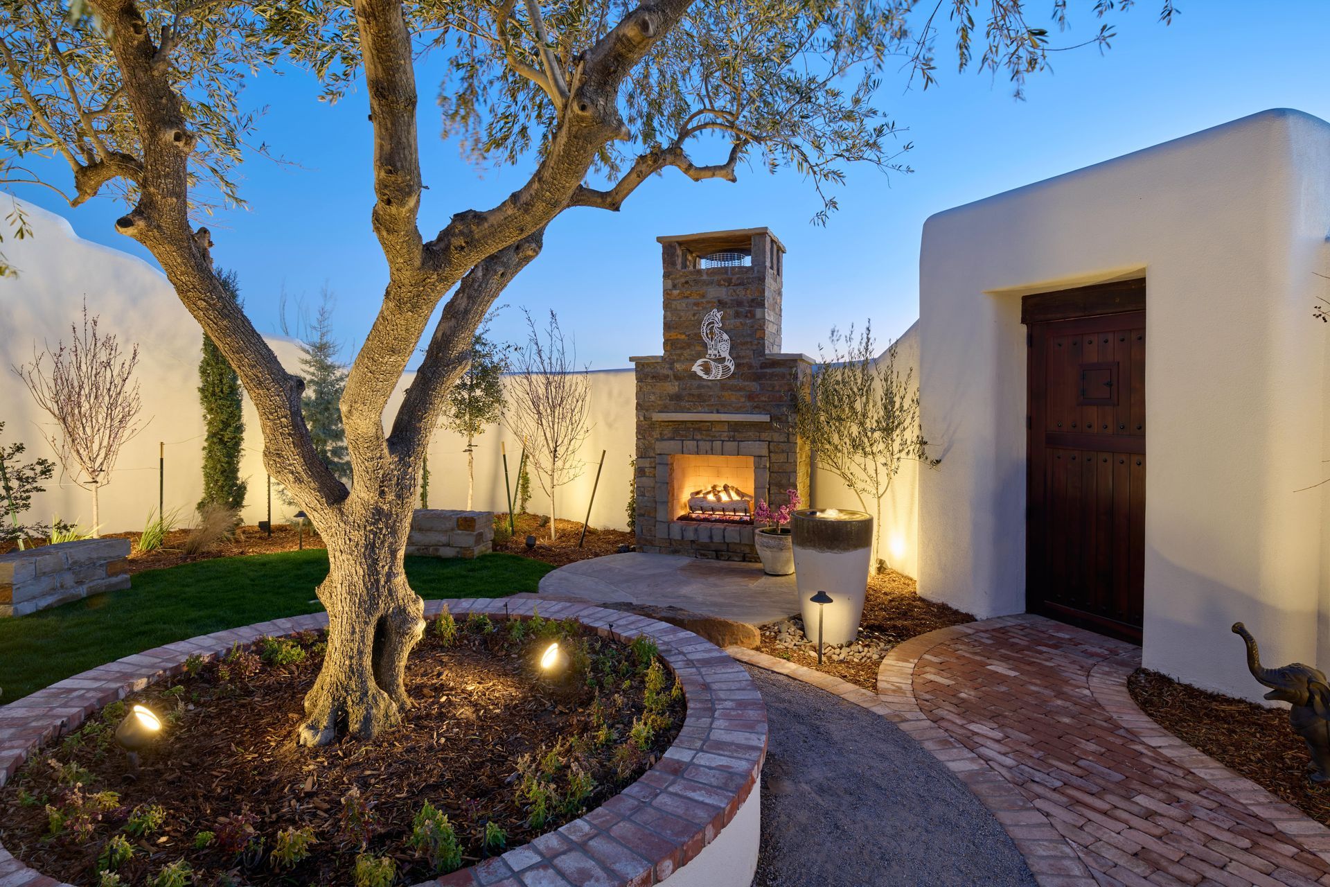 Backyard with olive tree, fireplace, and path lit by spotlights. Whitewashed walls and a wooden door.