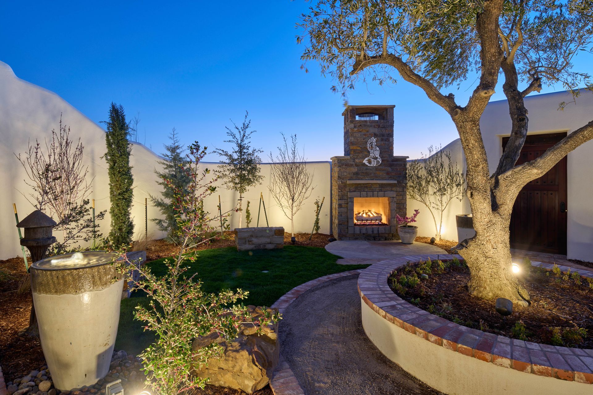Backyard courtyard with stone fireplace, fountain, tree, and pathway, illuminated at dusk.