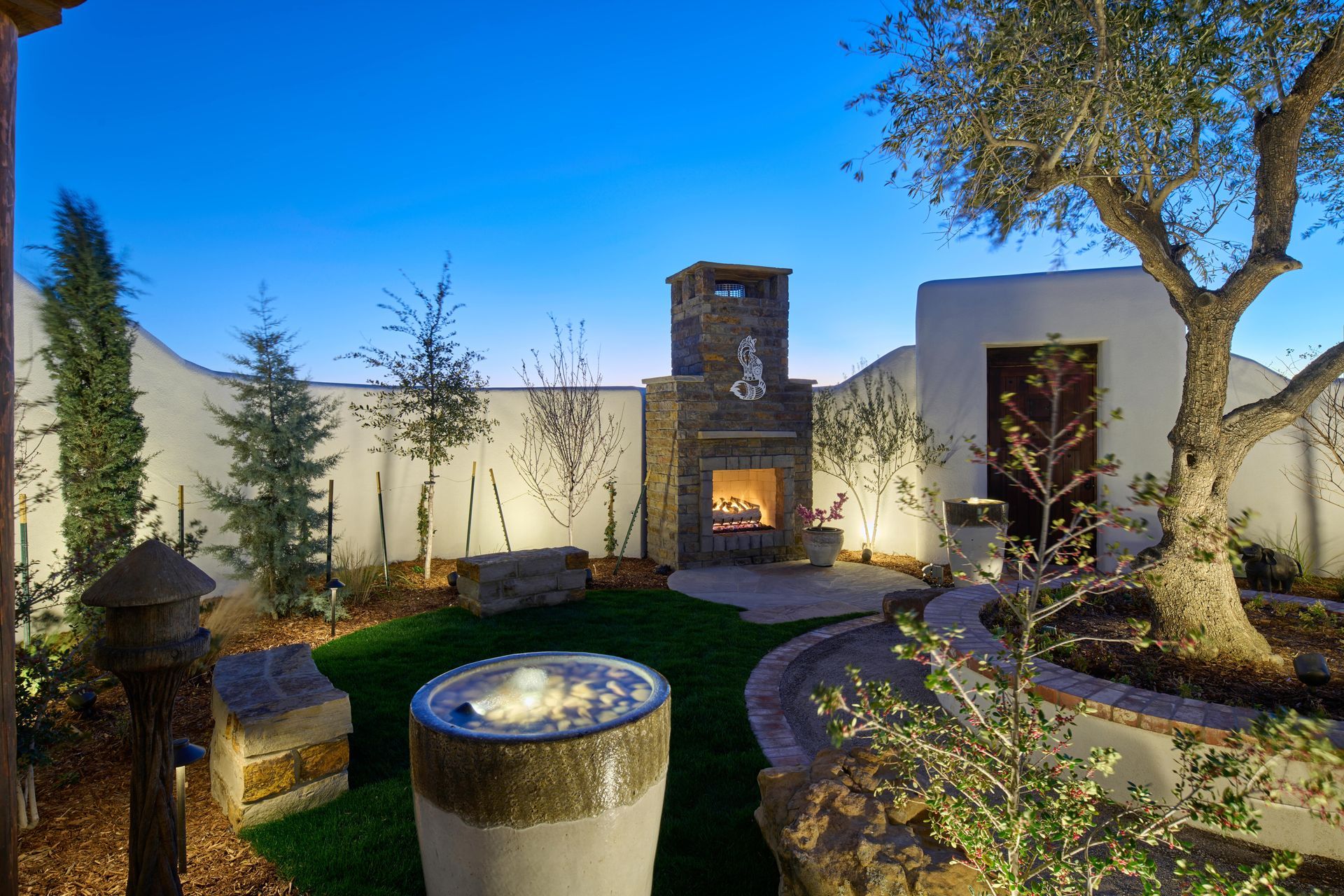 Outdoor patio at dusk with a fireplace, water feature, and mature tree.