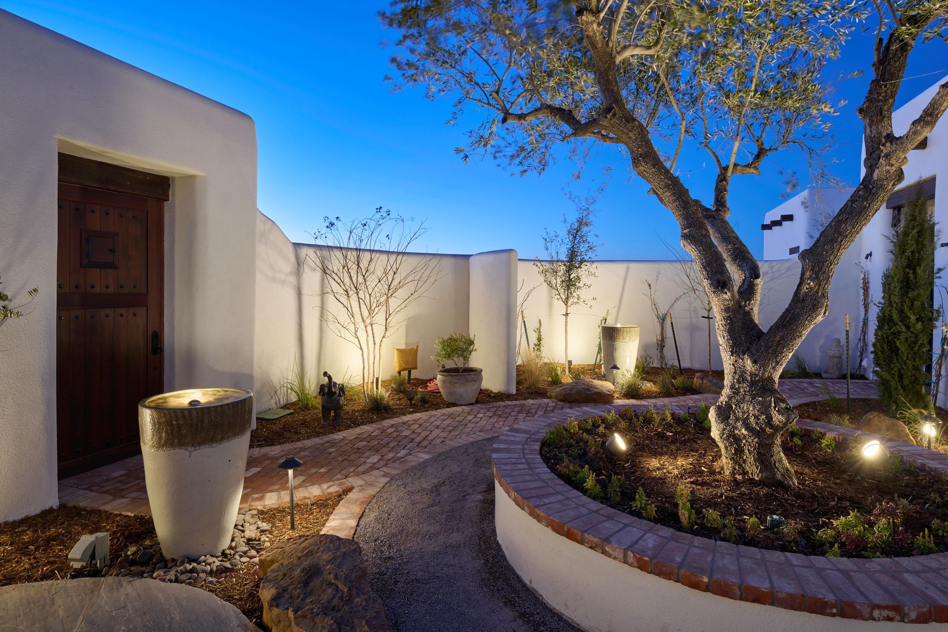 Courtyard with brick pathway, mature tree, and outdoor lighting against a white stucco wall at dusk.