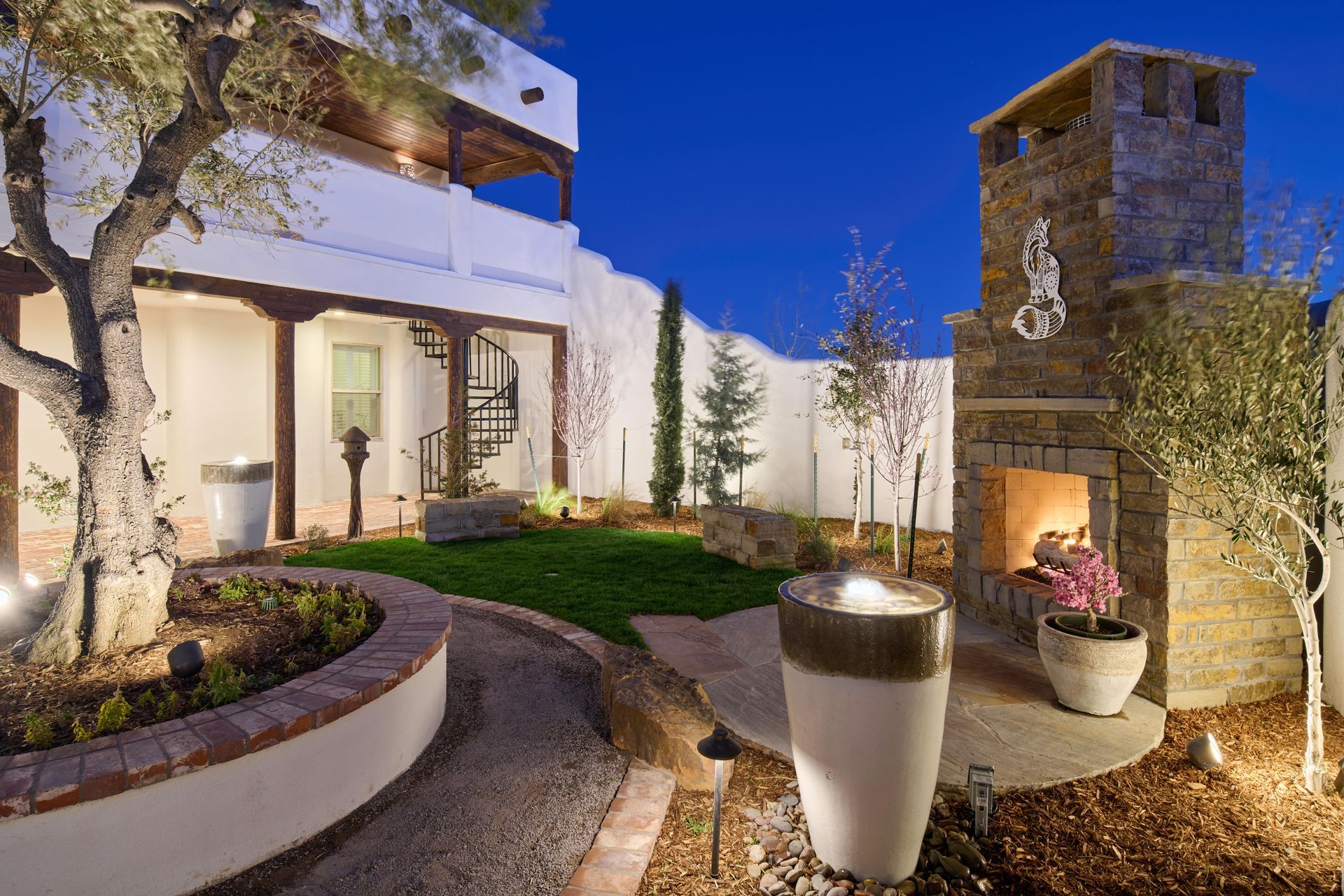 Courtyard with fireplace, trees, and architectural details at dusk; white walls, stone accents, and spiral staircase.