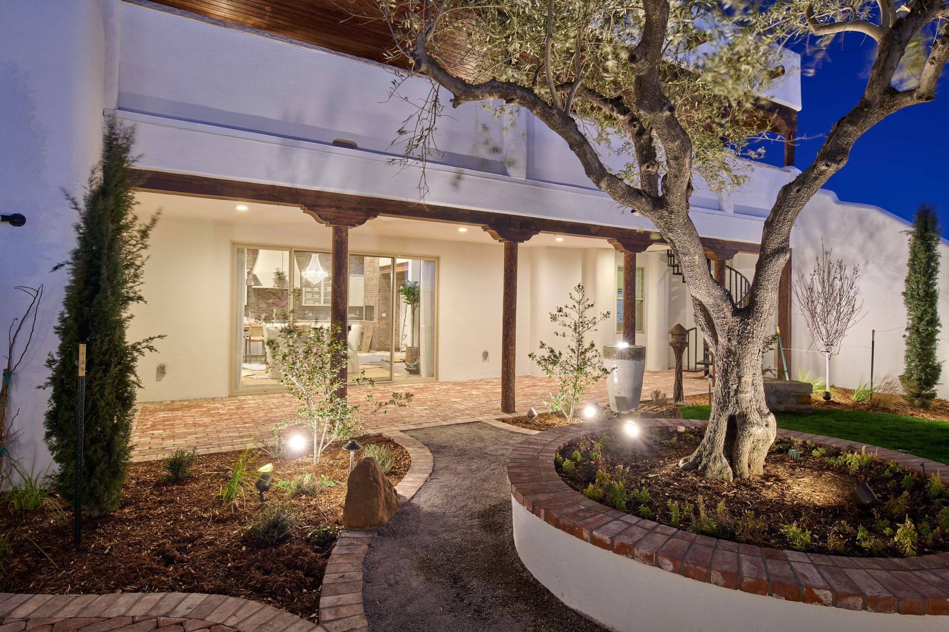 Exterior patio with mature tree, brick pathway, and white stucco building at dusk.