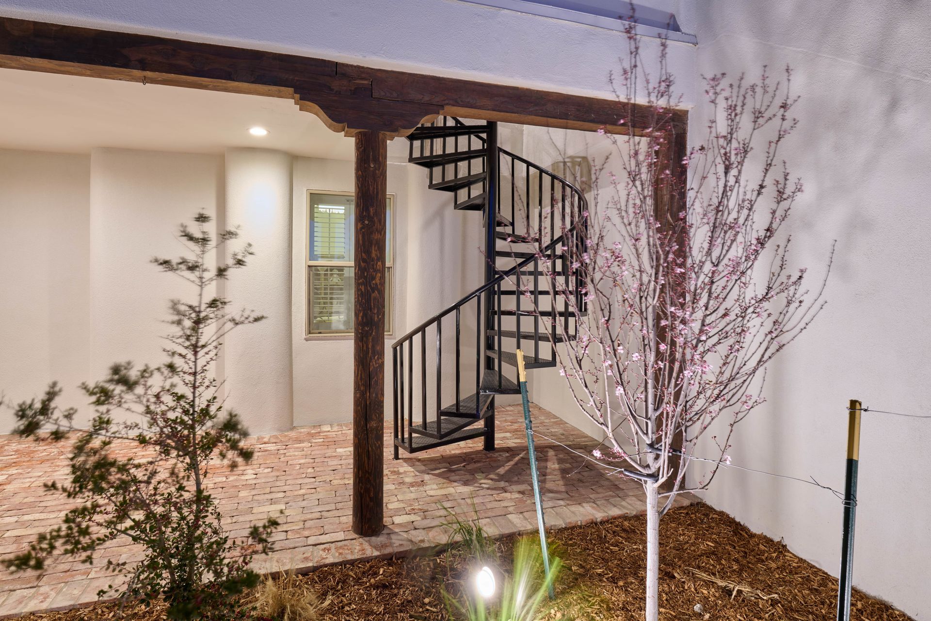Spiral staircase under a wooden beam, with brick floor and sparse landscaping.