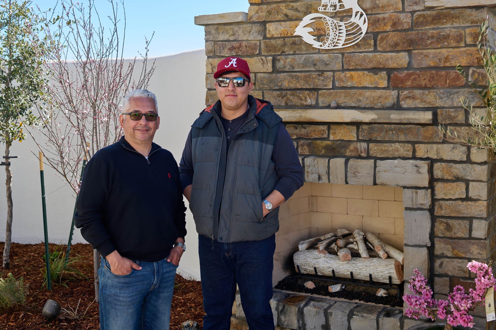 Two men standing in front of a stone fireplace. One wears a baseball cap and vest, the other, a sweater.