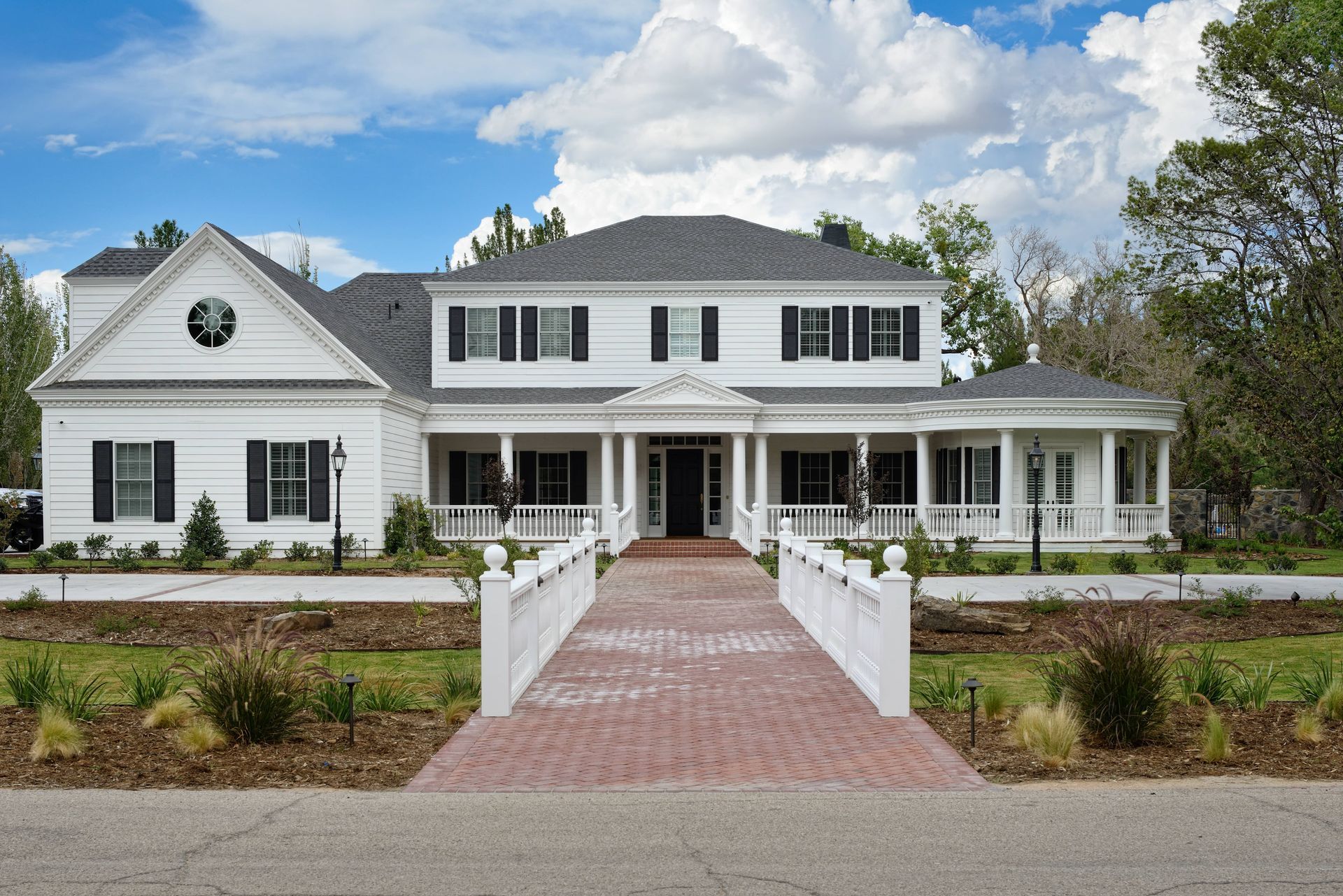 White two-story house with black shutters, columns, and a brick walkway under a blue sky.