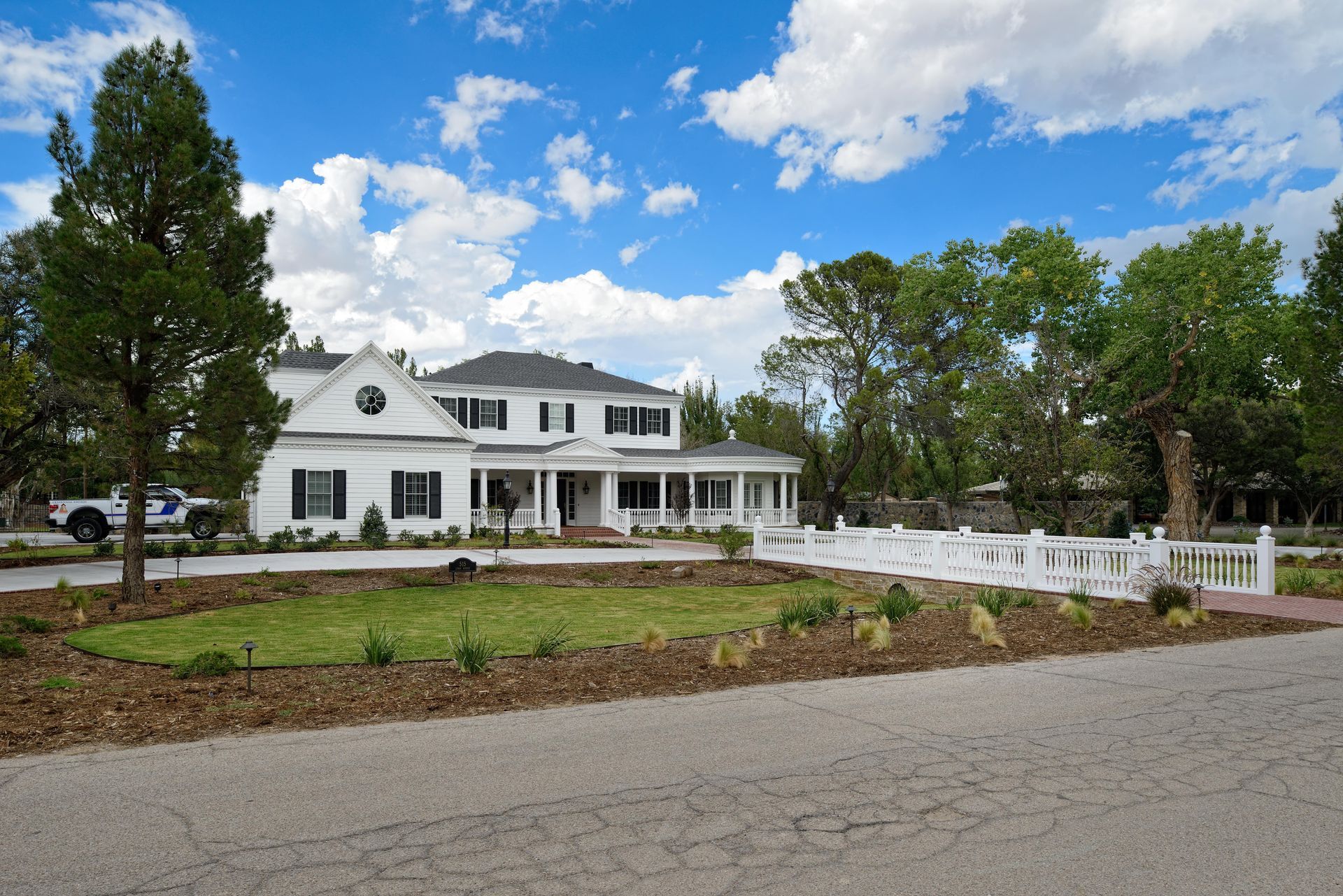 White two-story house with black shutters, wraparound porch, and picket fence, under a partly cloudy blue sky.