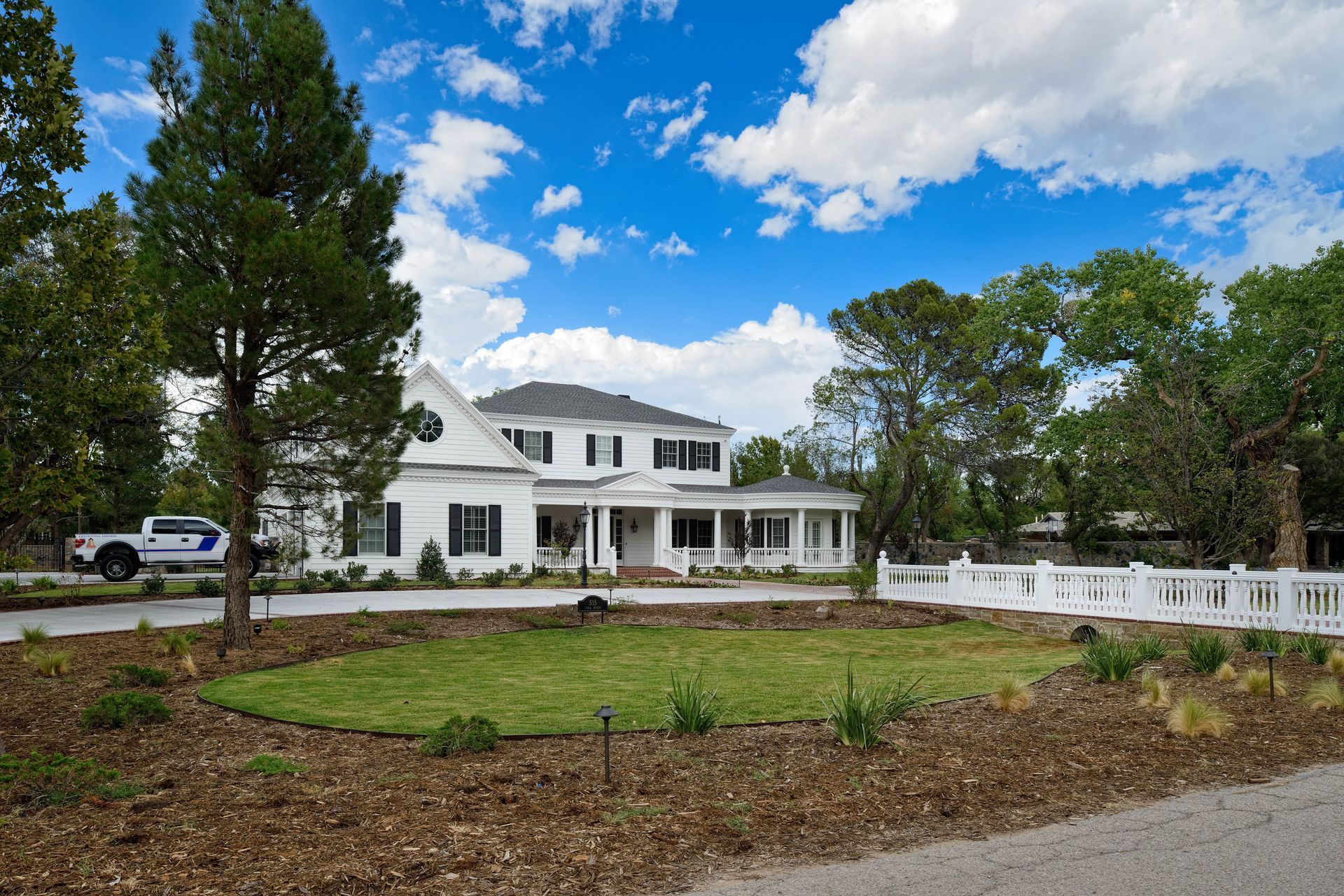White two-story house with front porch and black shutters, set on a green lawn with a white fence. Blue sky and trees.