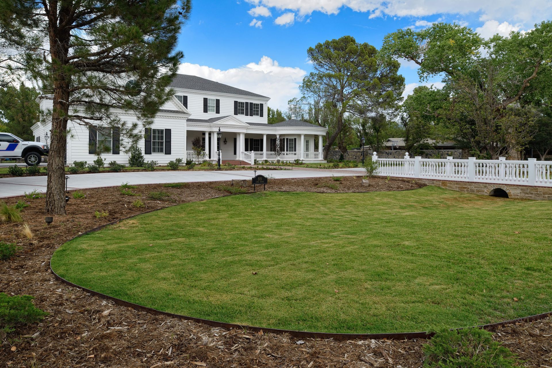 White house with a large green lawn, white picket fence, and surrounding trees under a cloudy blue sky.
