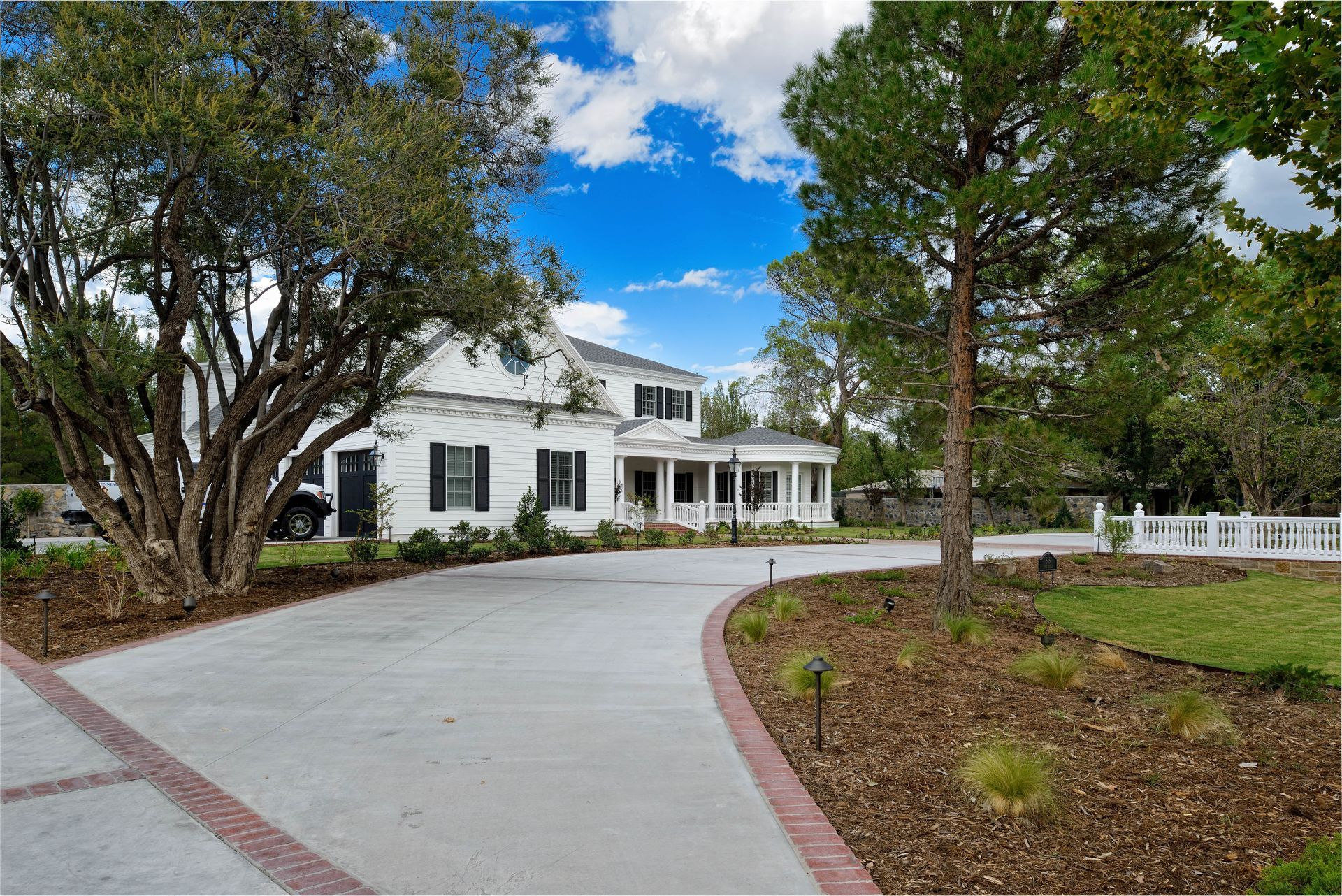 White two-story house with a long concrete driveway, bordered by landscaping and trees, under a blue sky.