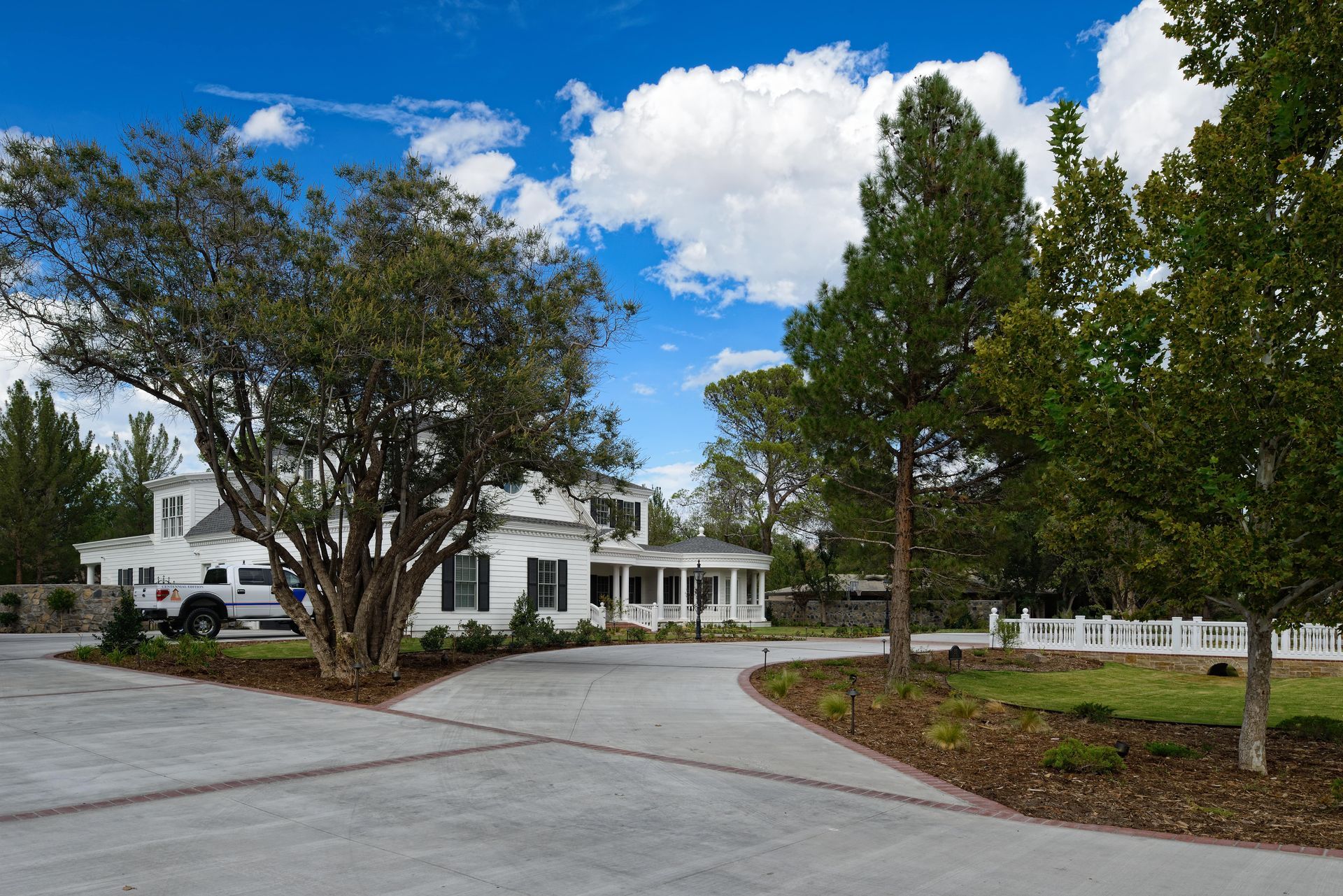 White house with long porch, driveway, and trees under a blue sky with clouds.