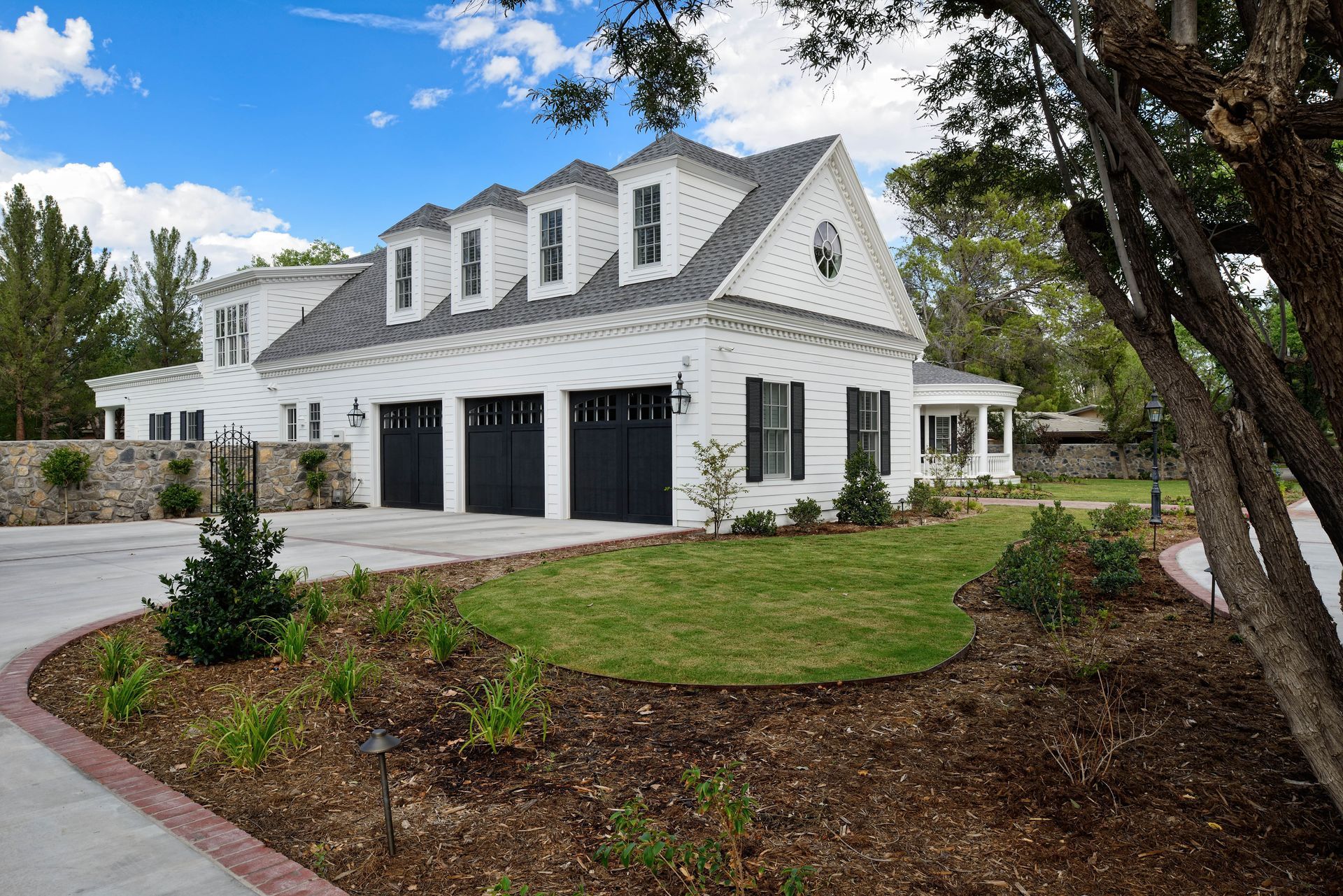 White house with black garage doors, dormers, and landscaping under a blue sky.