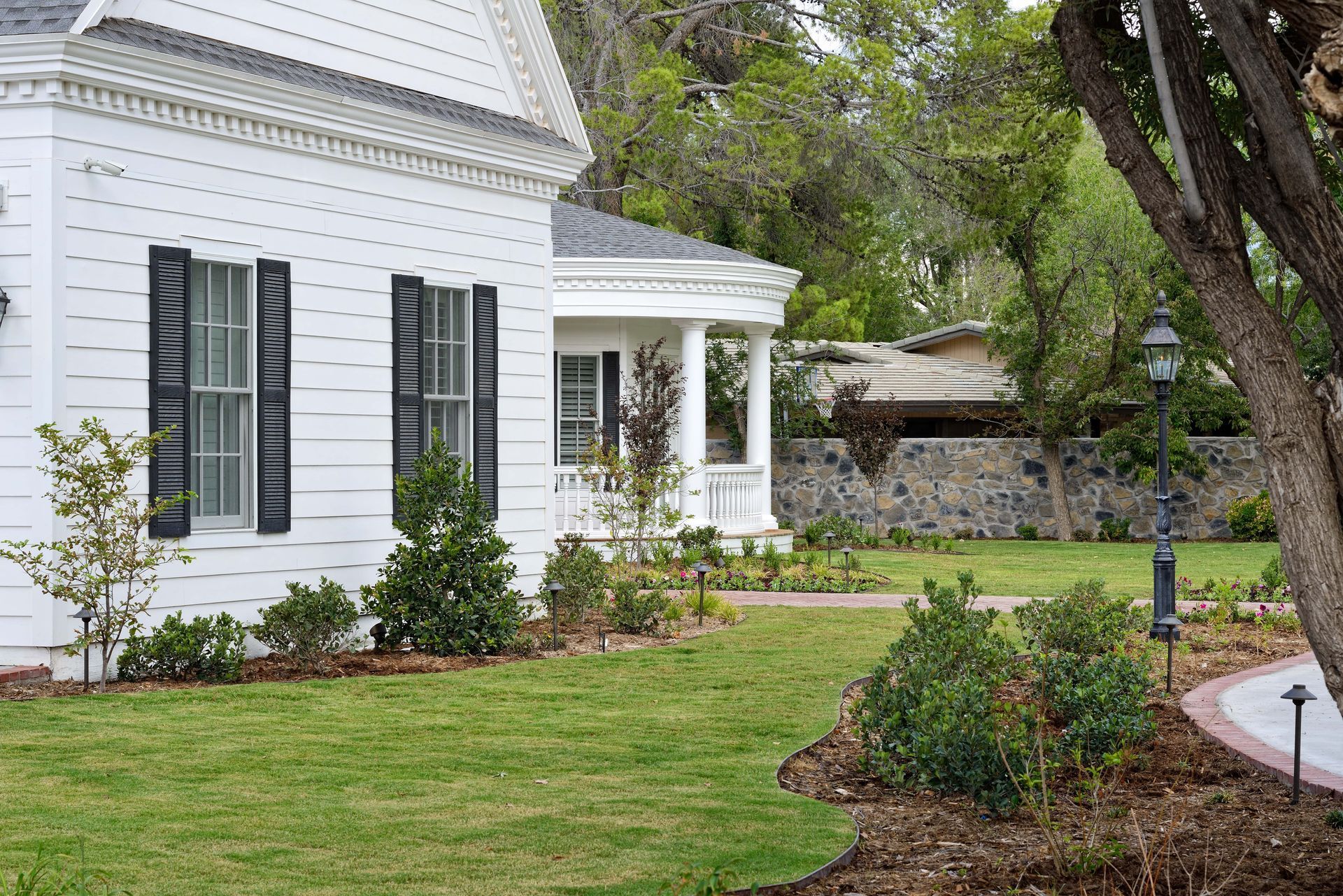White house with black shutters, green lawn, trees, and garden path.