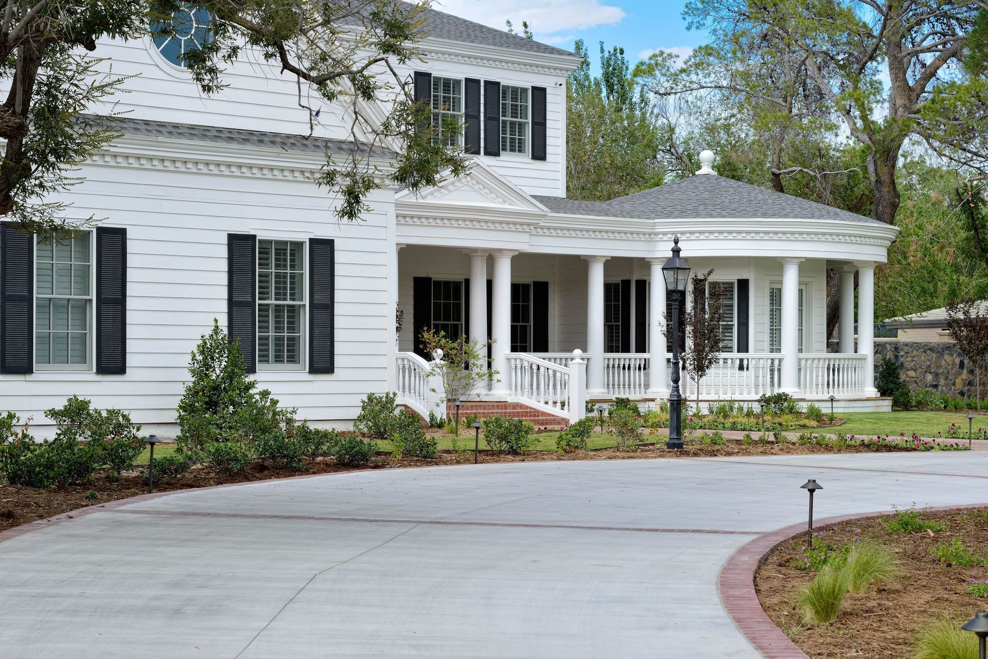 White colonial house with black shutters and a wraparound porch. Concrete driveway.