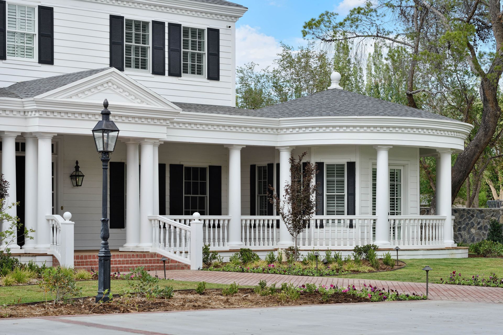 White colonial house with black shutters, wraparound porch, and a brick pathway.