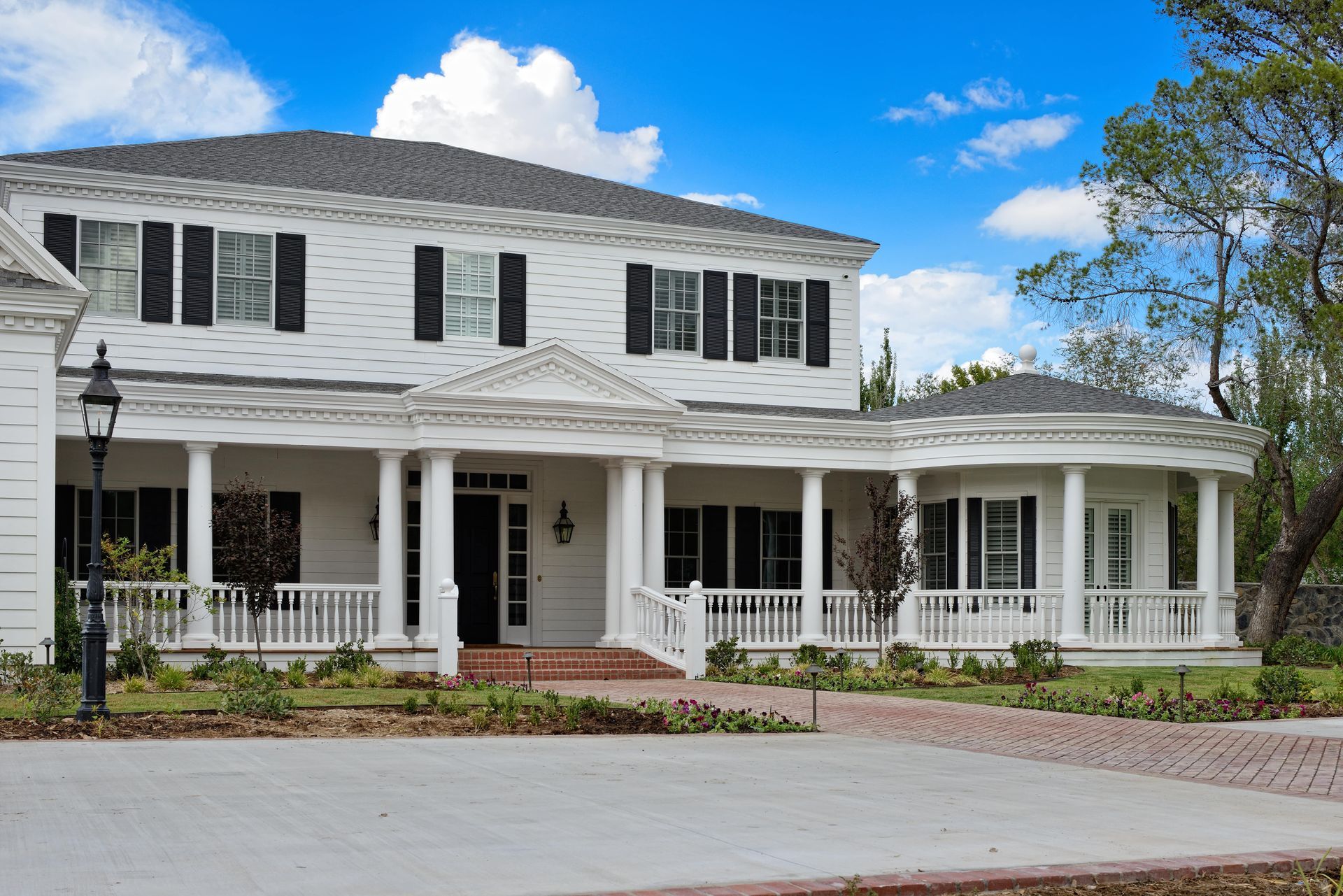 White two-story house with black shutters, large porch, brick driveway, and blue sky.