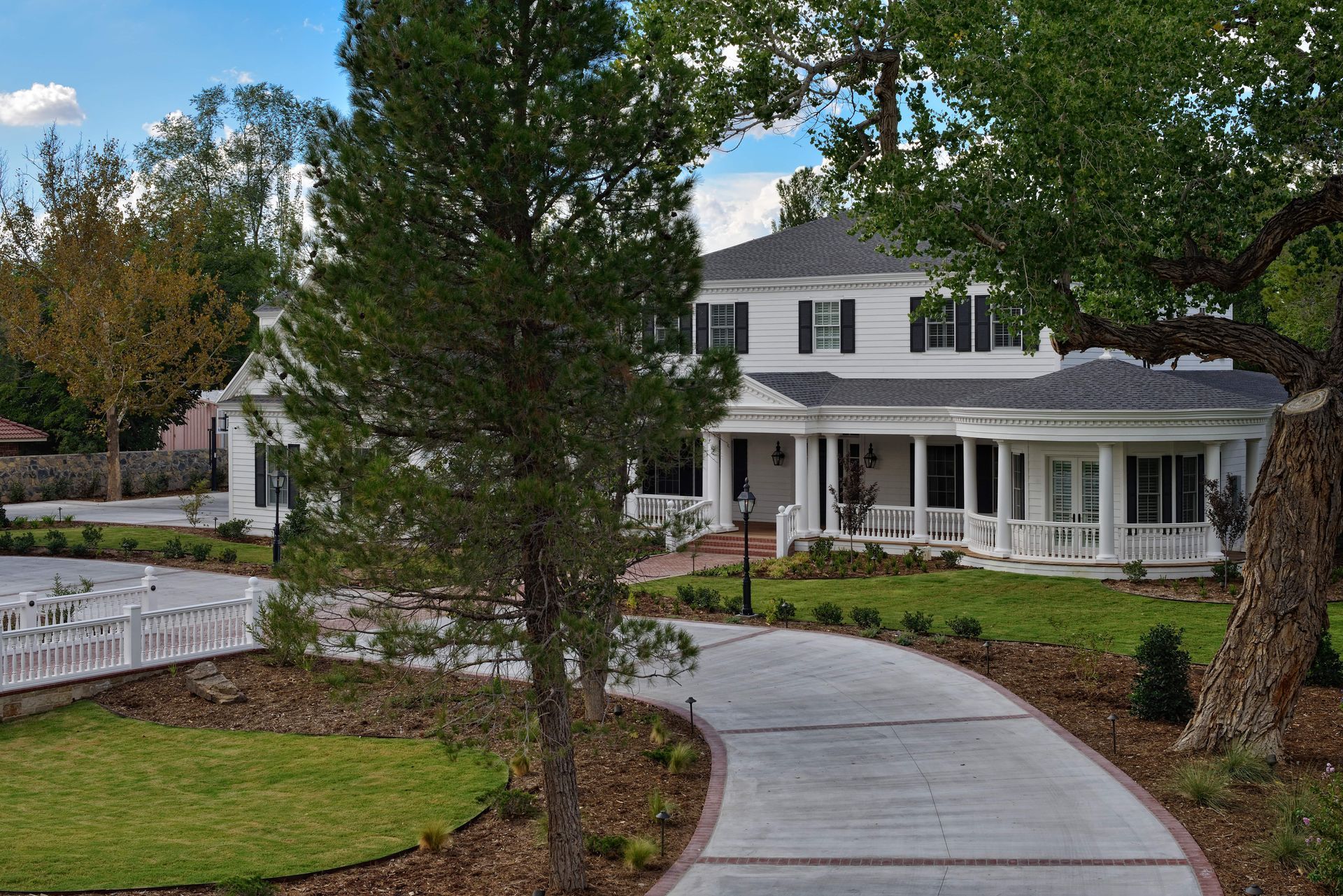 White colonial house with wraparound porch and curved driveway. Lush green lawn with trees and blue sky.