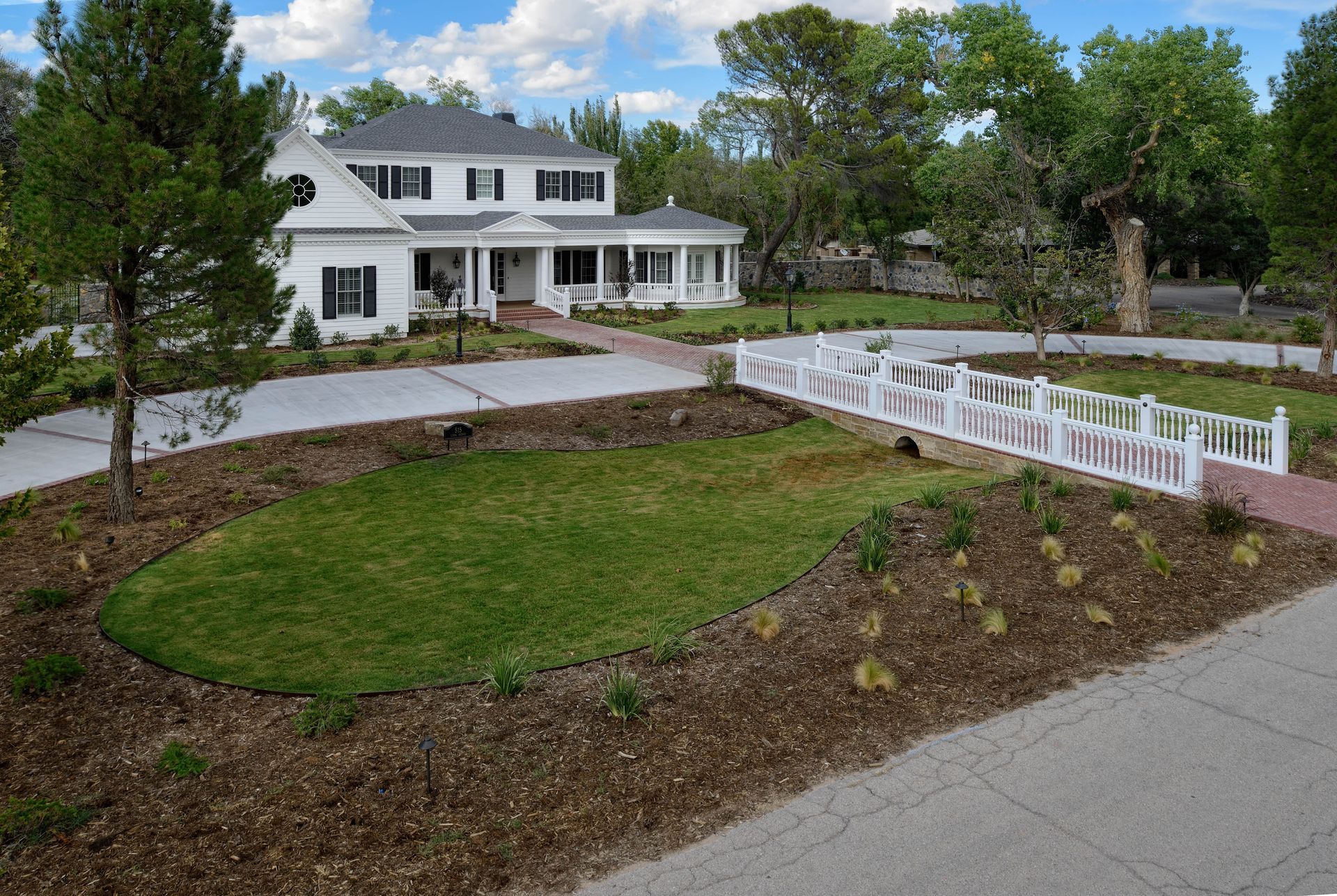 White two-story house with black shutters, long driveway, and a white bridge over a small grassy area.
