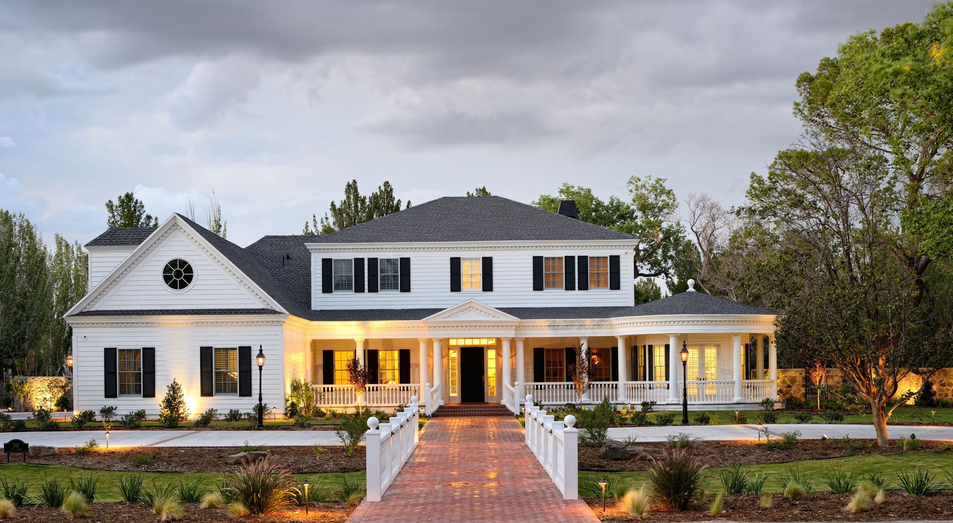 White two-story house with black shutters and a wraparound porch, brick path leading to the front door.