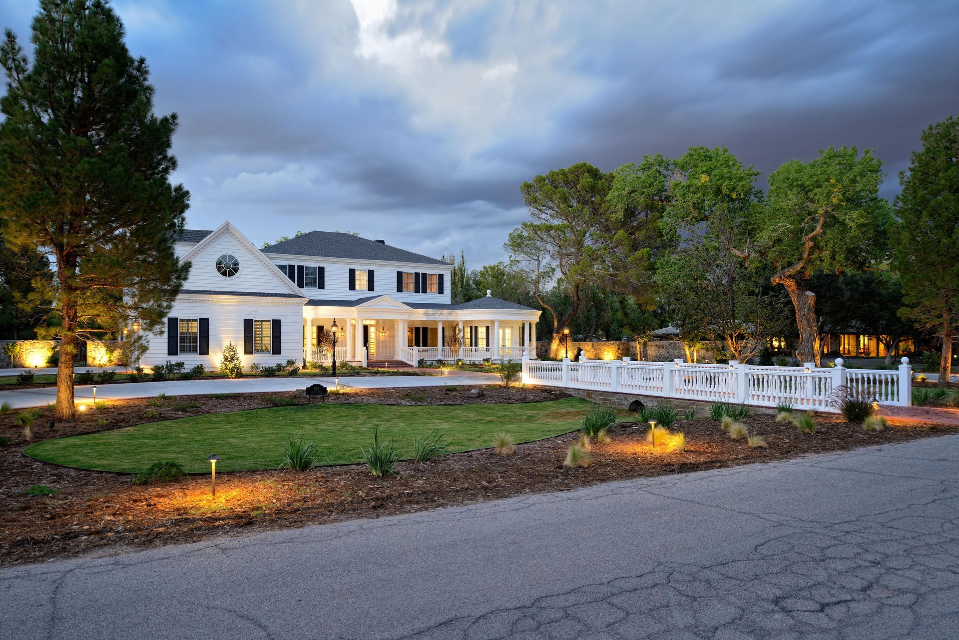White two-story house with black shutters, white porch, and fenced yard at dusk under cloudy sky.