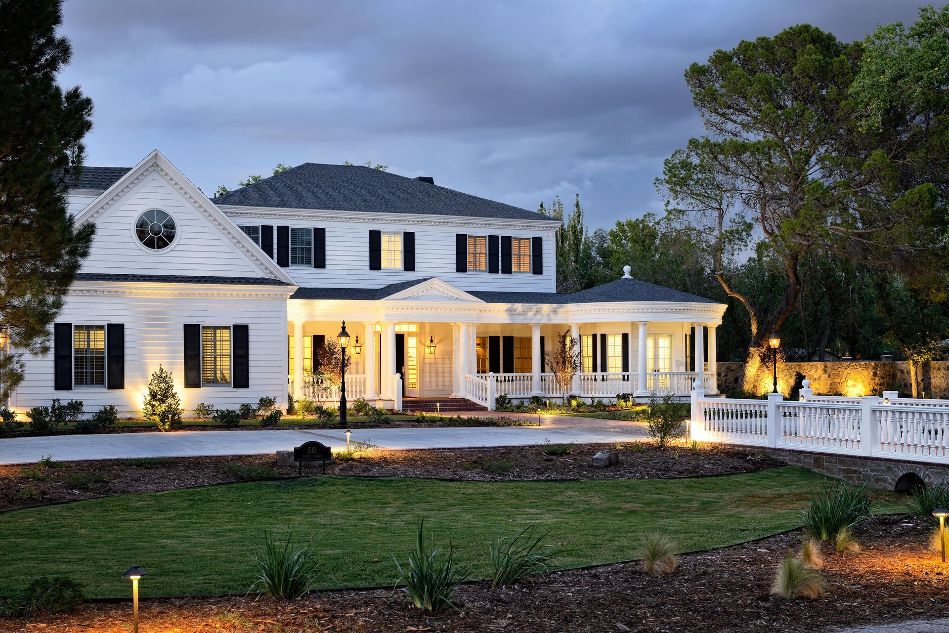 White two-story house with porch and black shutters at dusk; lit landscaping and driveway.