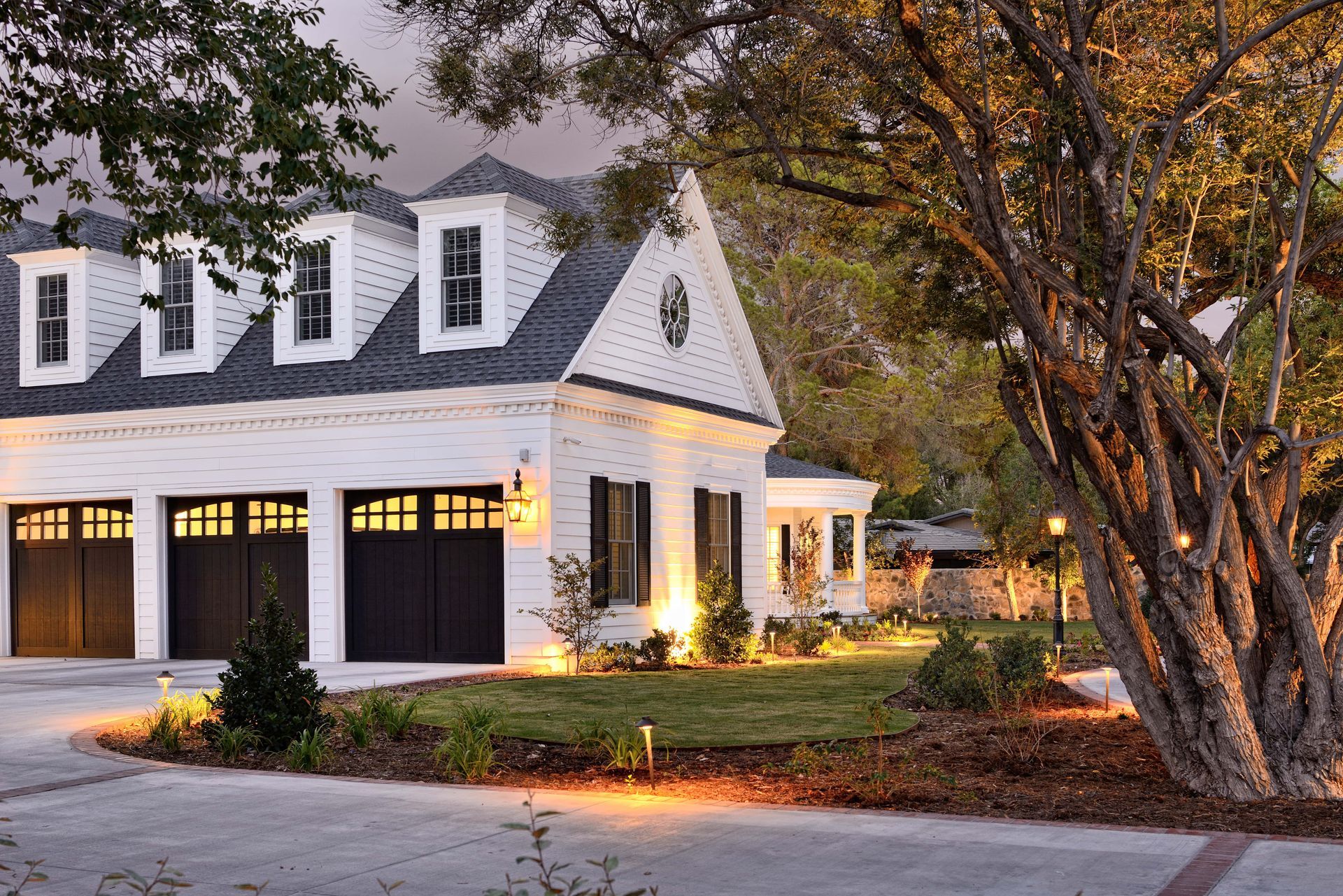 White two-story house with black garage doors, dormer windows, and a tree in the yard.
