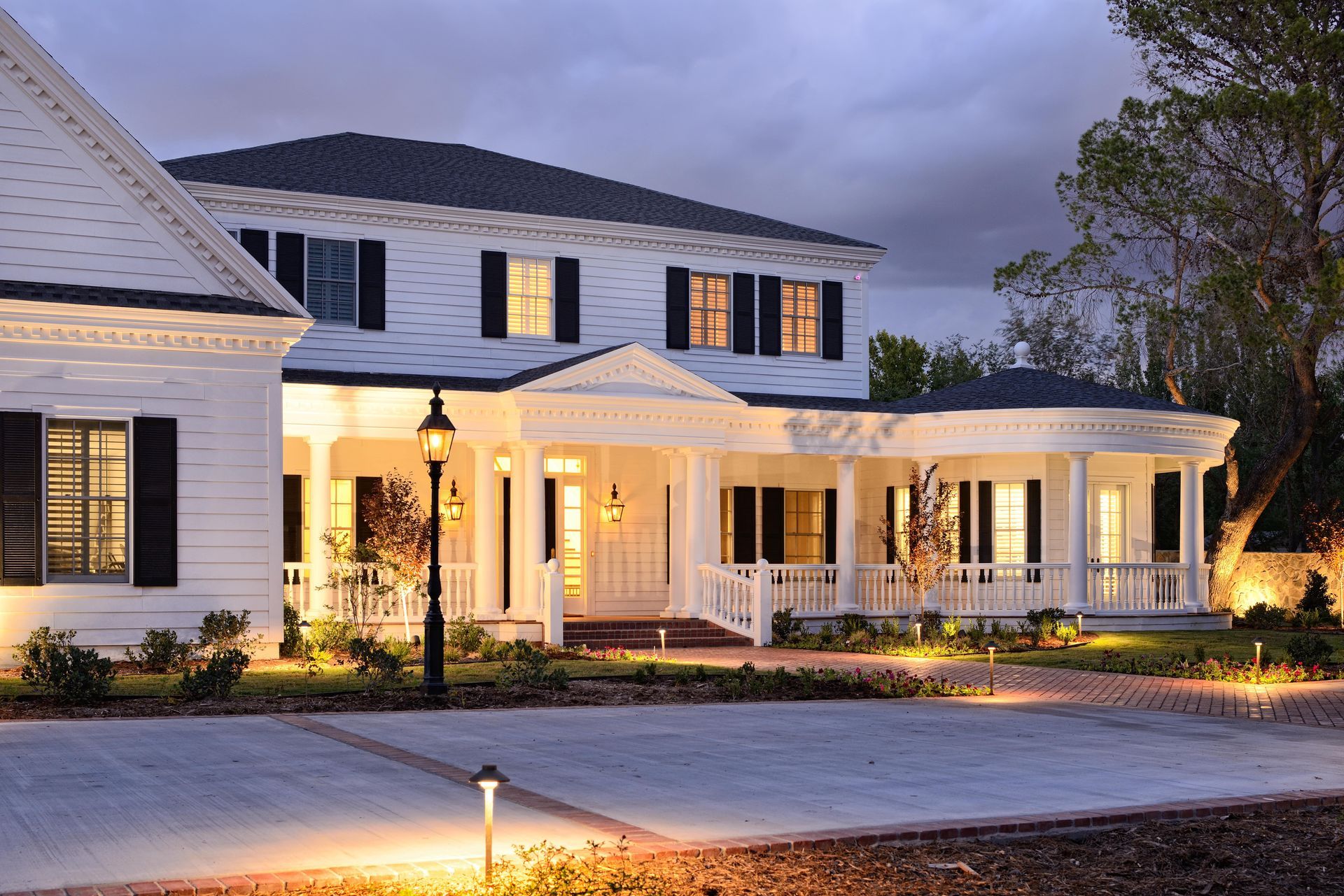 White two-story house with porch and black shutters, lit by outdoor lights. Evening setting.