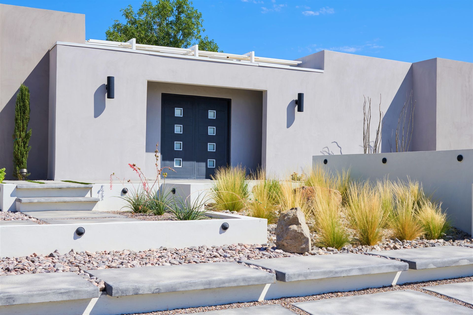 Modern home exterior with gray stucco, steps, landscaping, and a dark door.