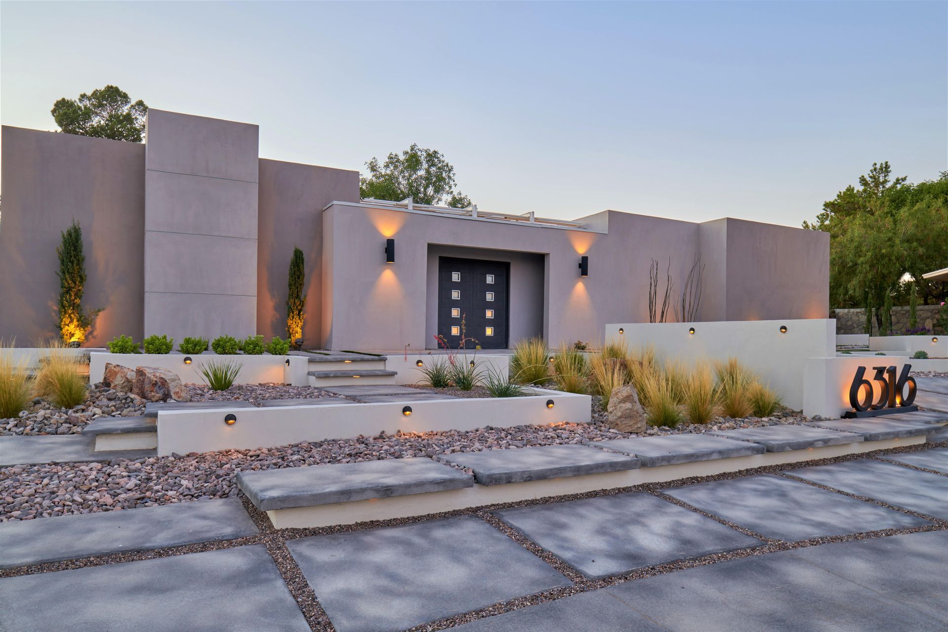 Modern gray home exterior with illuminated entry, stone landscaping, and address numbers.