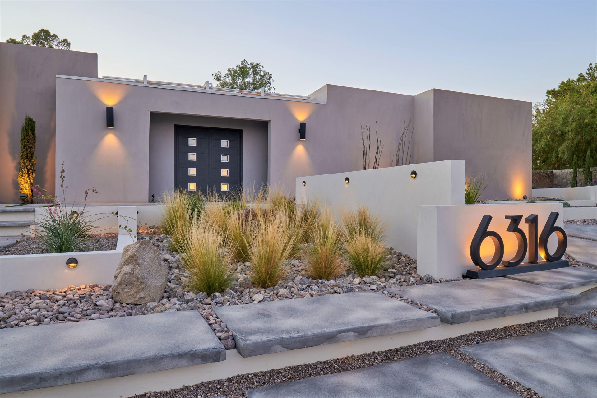 Modern home exterior with illuminated address numbers, gray concrete, and desert landscaping.