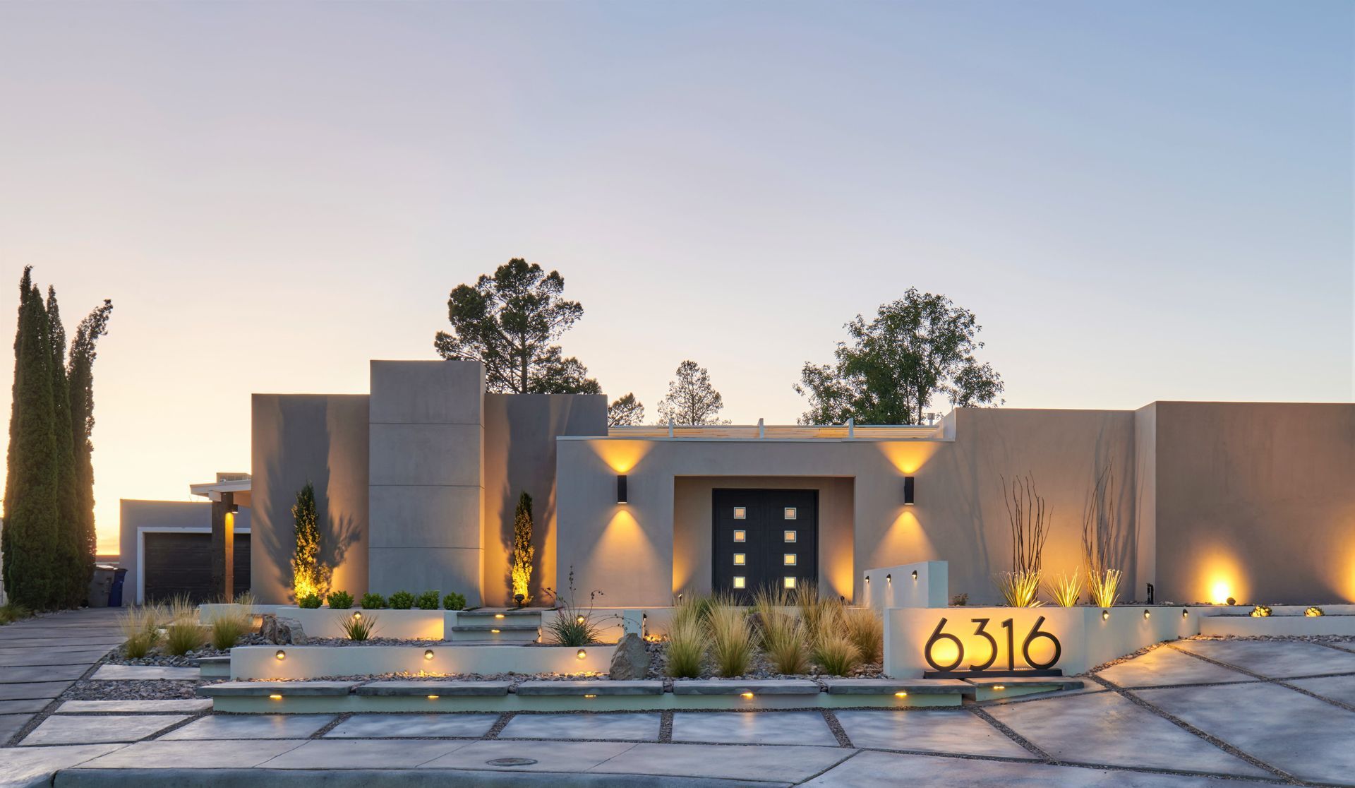 Modern, beige house with illuminated address numbers, landscaping, and a central entrance at dusk.