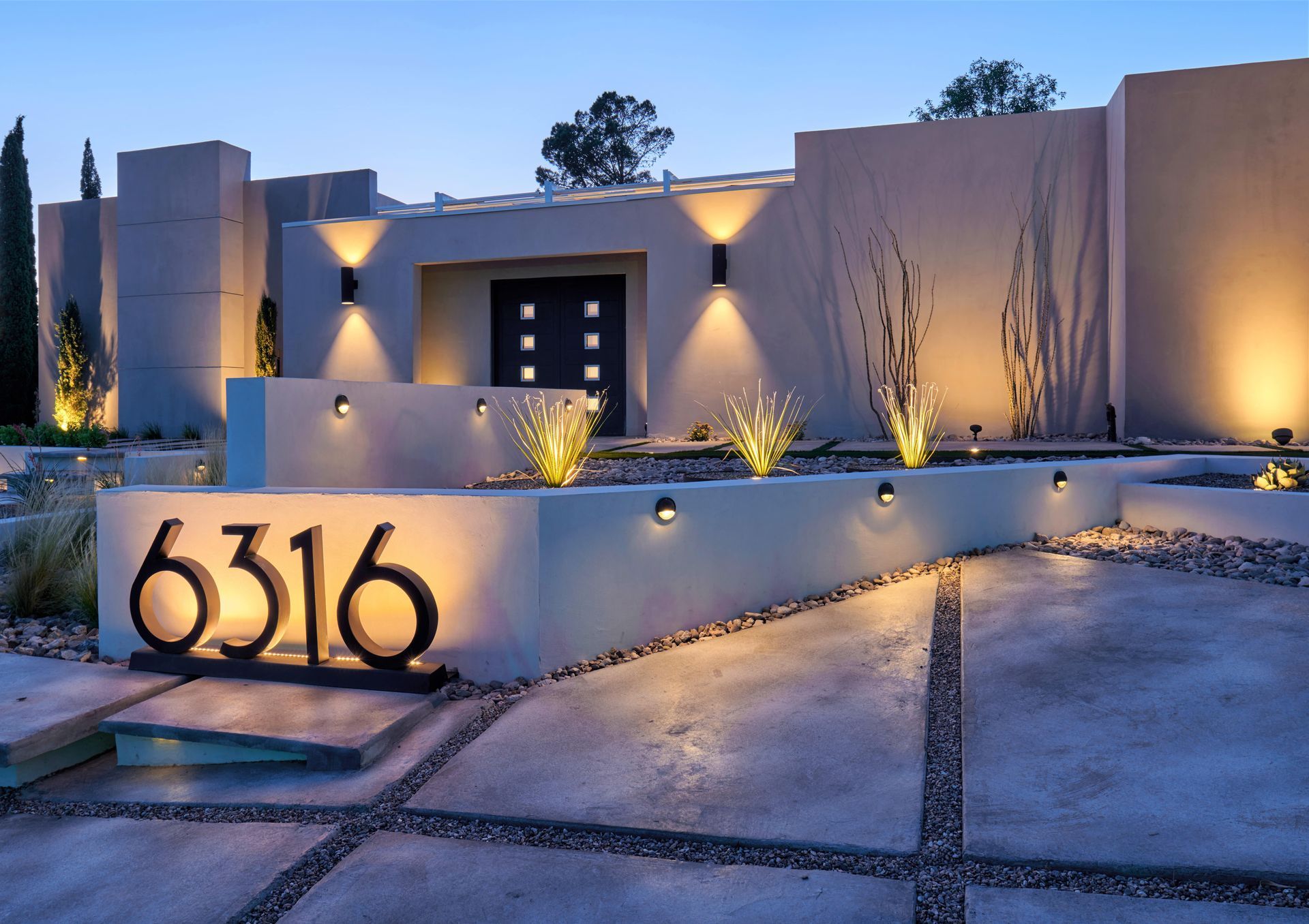 Modern home exterior with illuminated address numbers, concrete pathway, and landscaping at dusk.