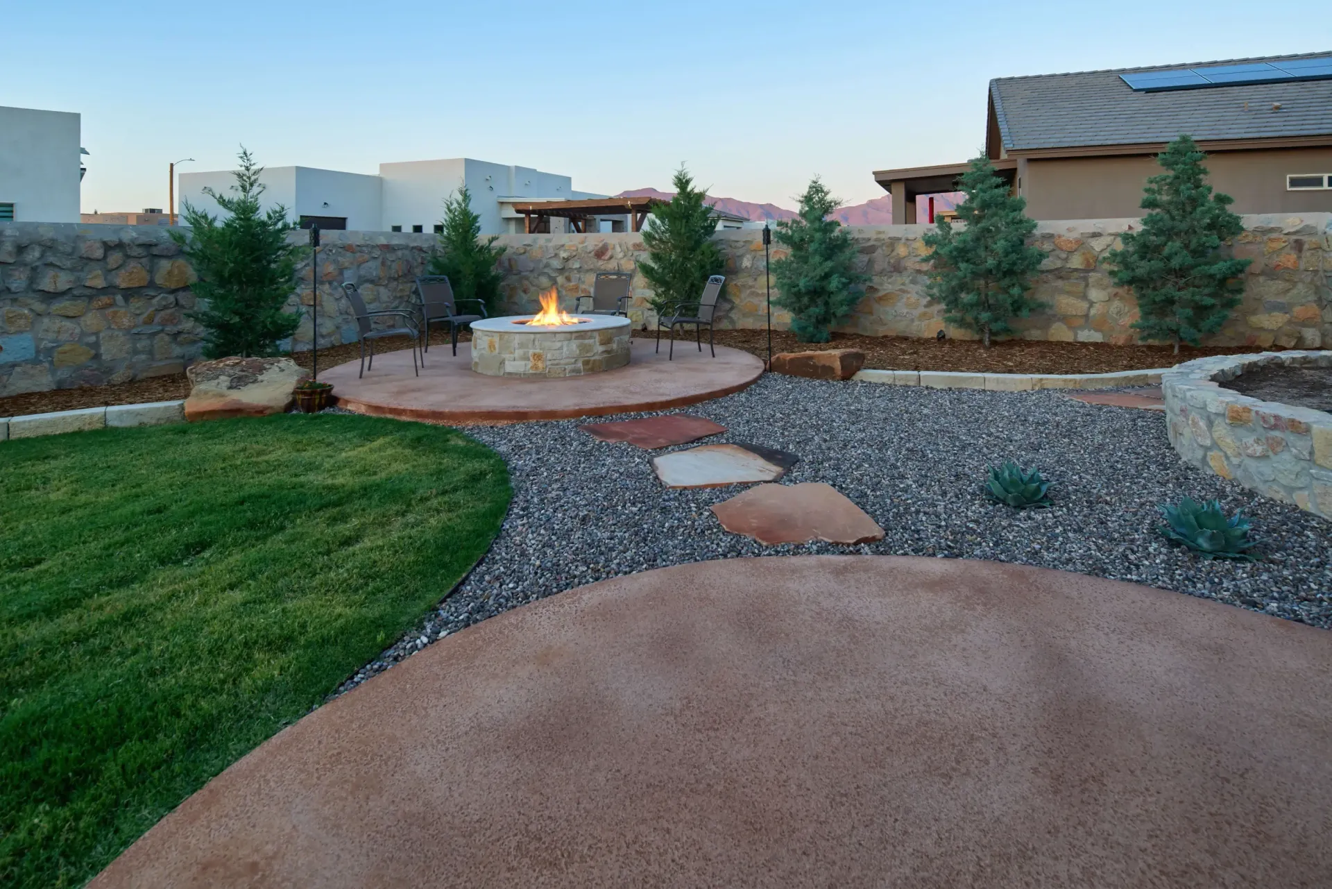 Backyard with fire pit on a circular patio, gravel, and grass. Trees and a stone wall backdrop.