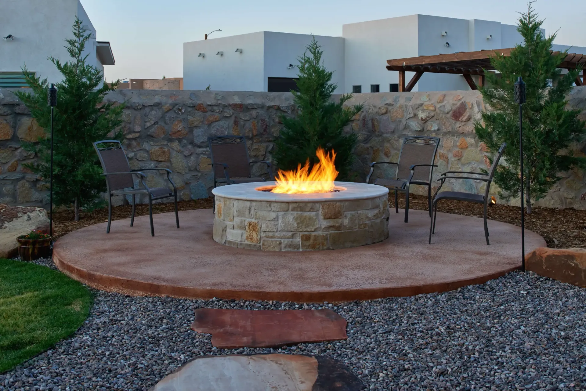 Stone fire pit with flames, chairs, and surrounding landscaping.