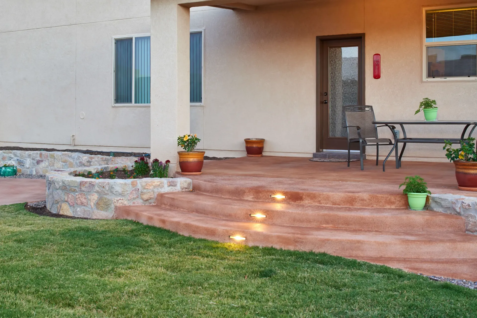 Backyard patio with steps, potted plants, and built-in lights, concrete and grass.