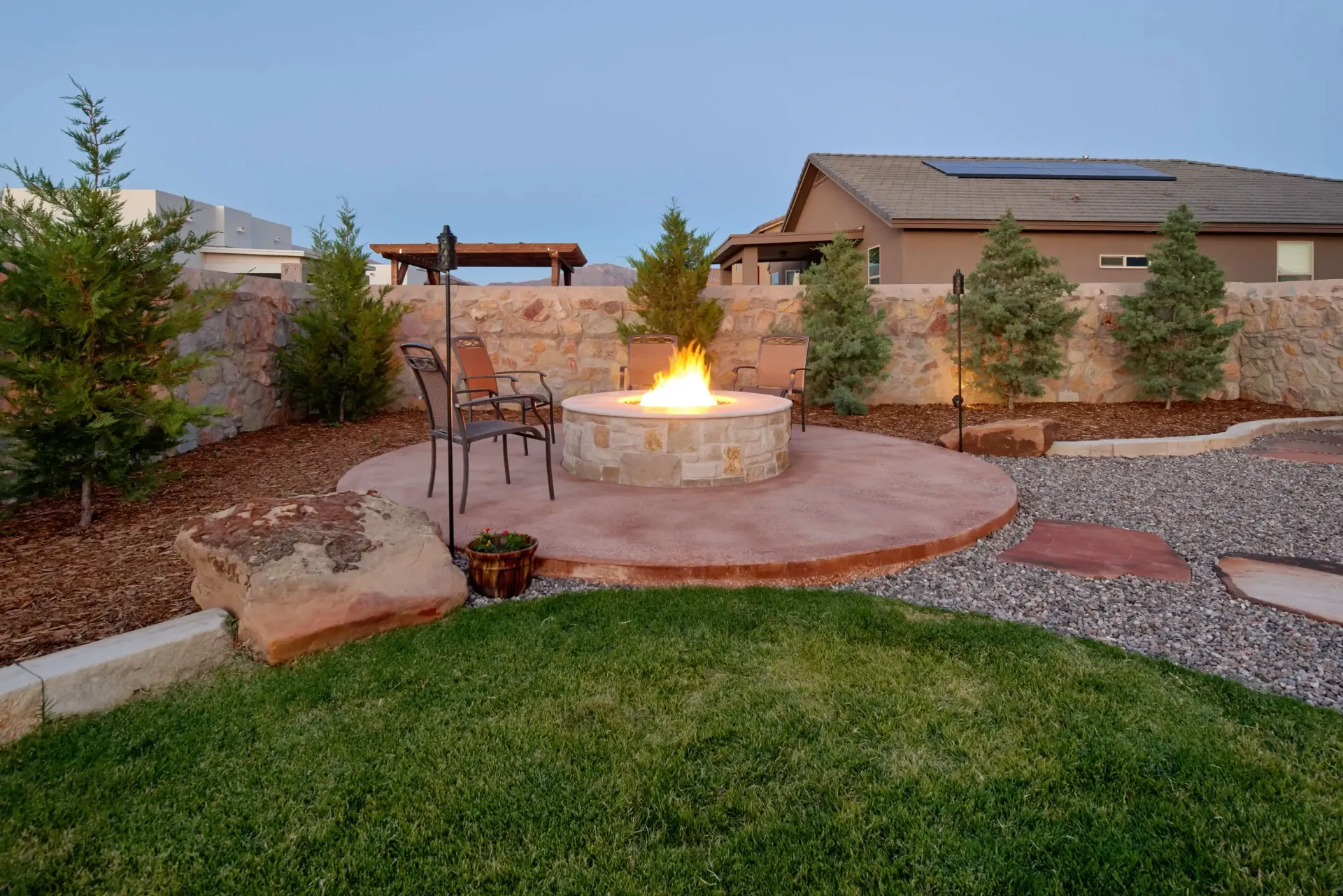 Backyard with a stone fire pit, chairs, and landscaping; evening scene.