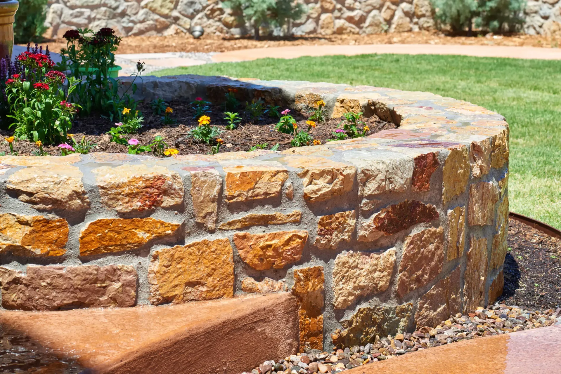 Curved stone flower bed filled with flowers, brown and gold stones, green grass.