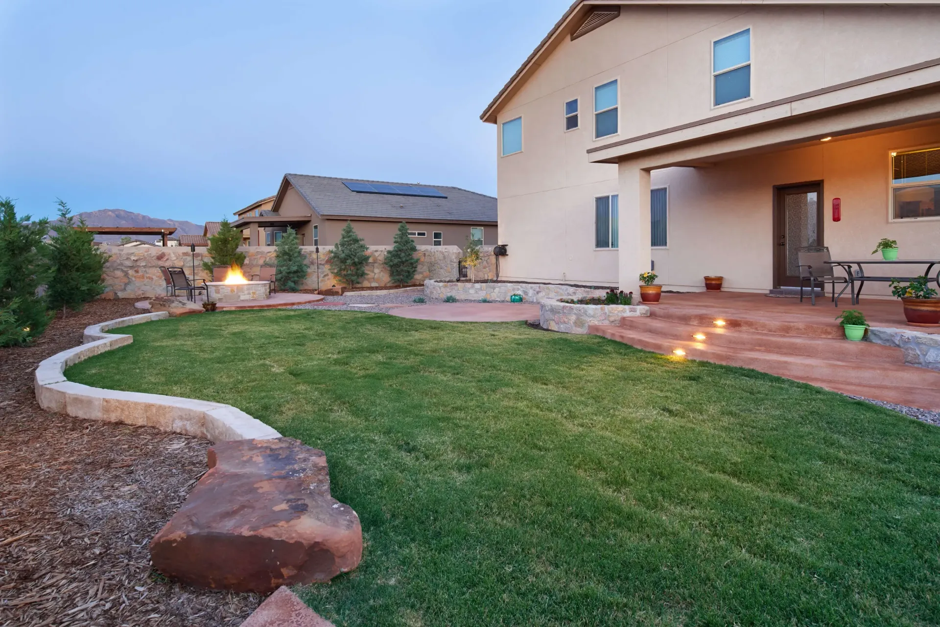 Backyard with green grass, stone retaining walls, fire pit, and a covered patio, in the evening.