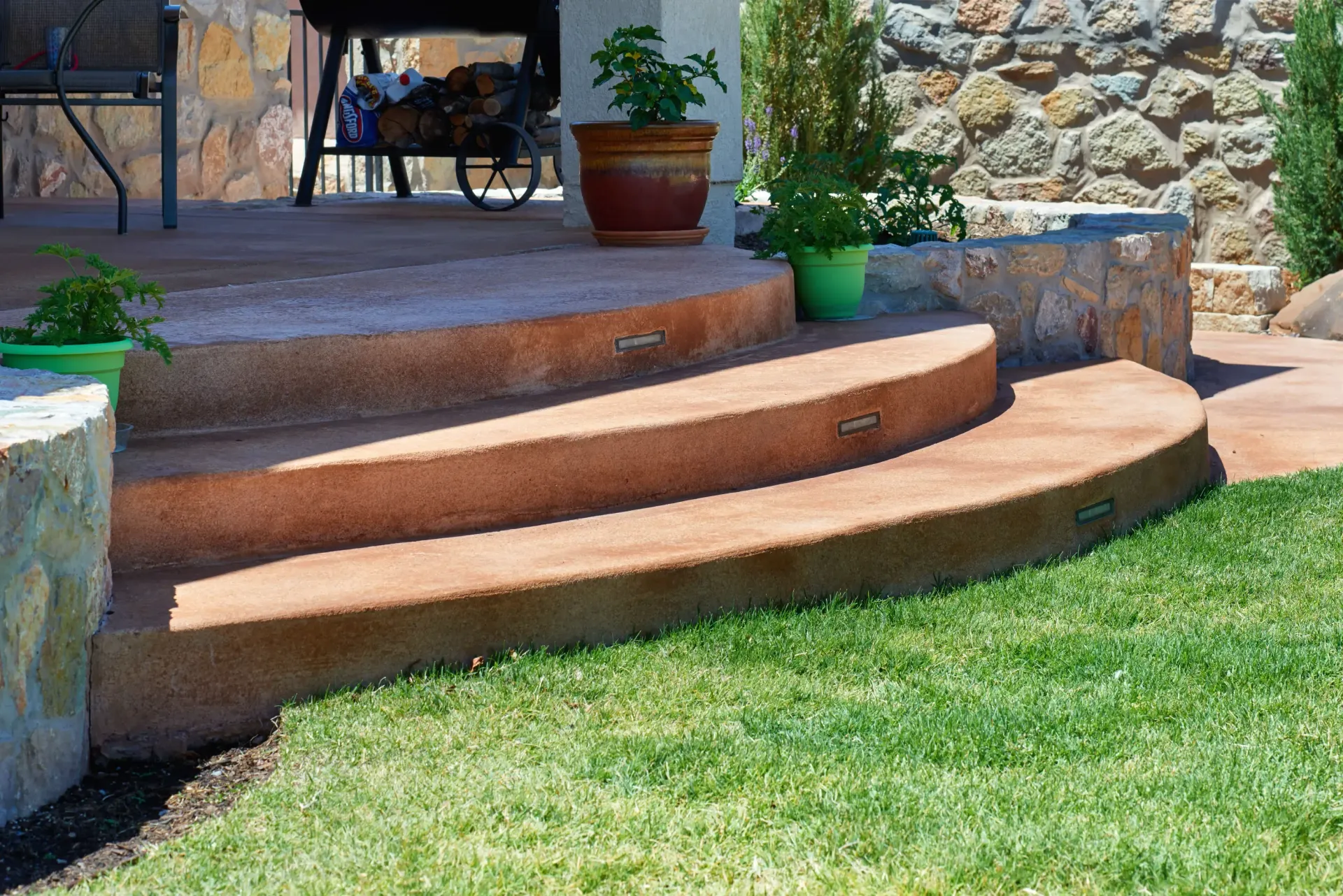 Curved, reddish-brown concrete steps lead to a patio, with potted plants nearby. Green lawn in front.
