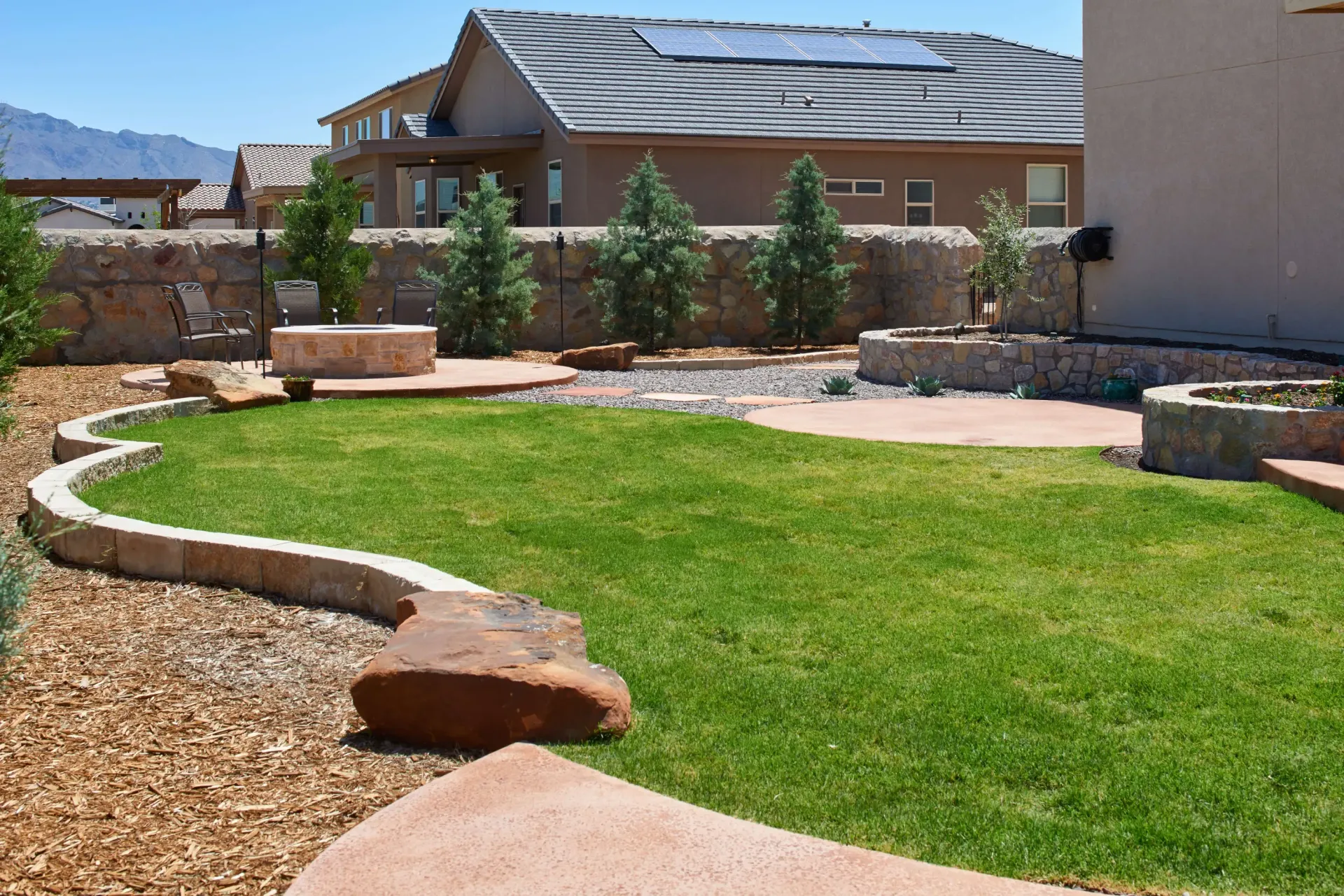 Backyard with green grass, stone patio, fire pits, and surrounding landscaping. Brown tones.