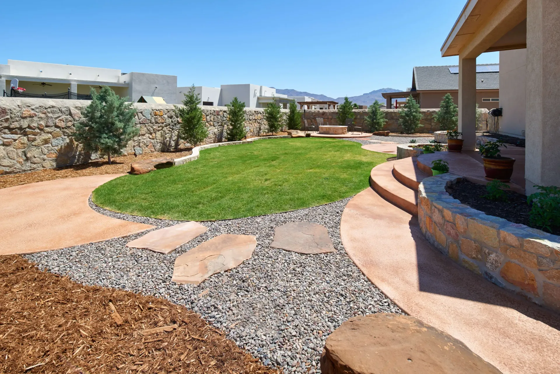 Backyard with grass, stone path, and shrubs against a blue sky, connecting to a home.