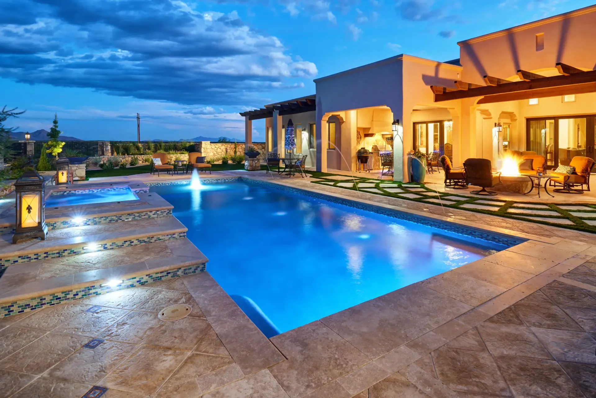 Pool and patio at dusk with illuminated pool, stone deck, and stucco home.