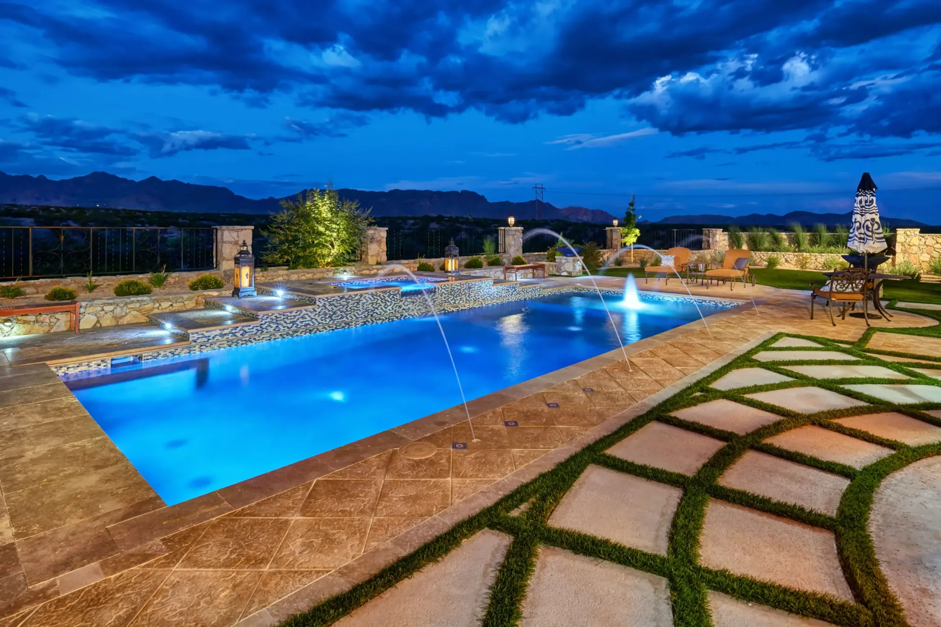 Nighttime view of a luxurious swimming pool with illuminated fountains, stone patio, and distant mountains under a cloudy blue sky.