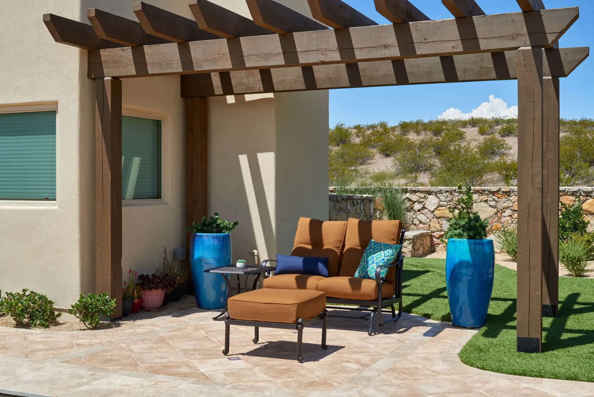 Patio with brown pergola, seating, and blue potted plants. Sunny day outdoors.