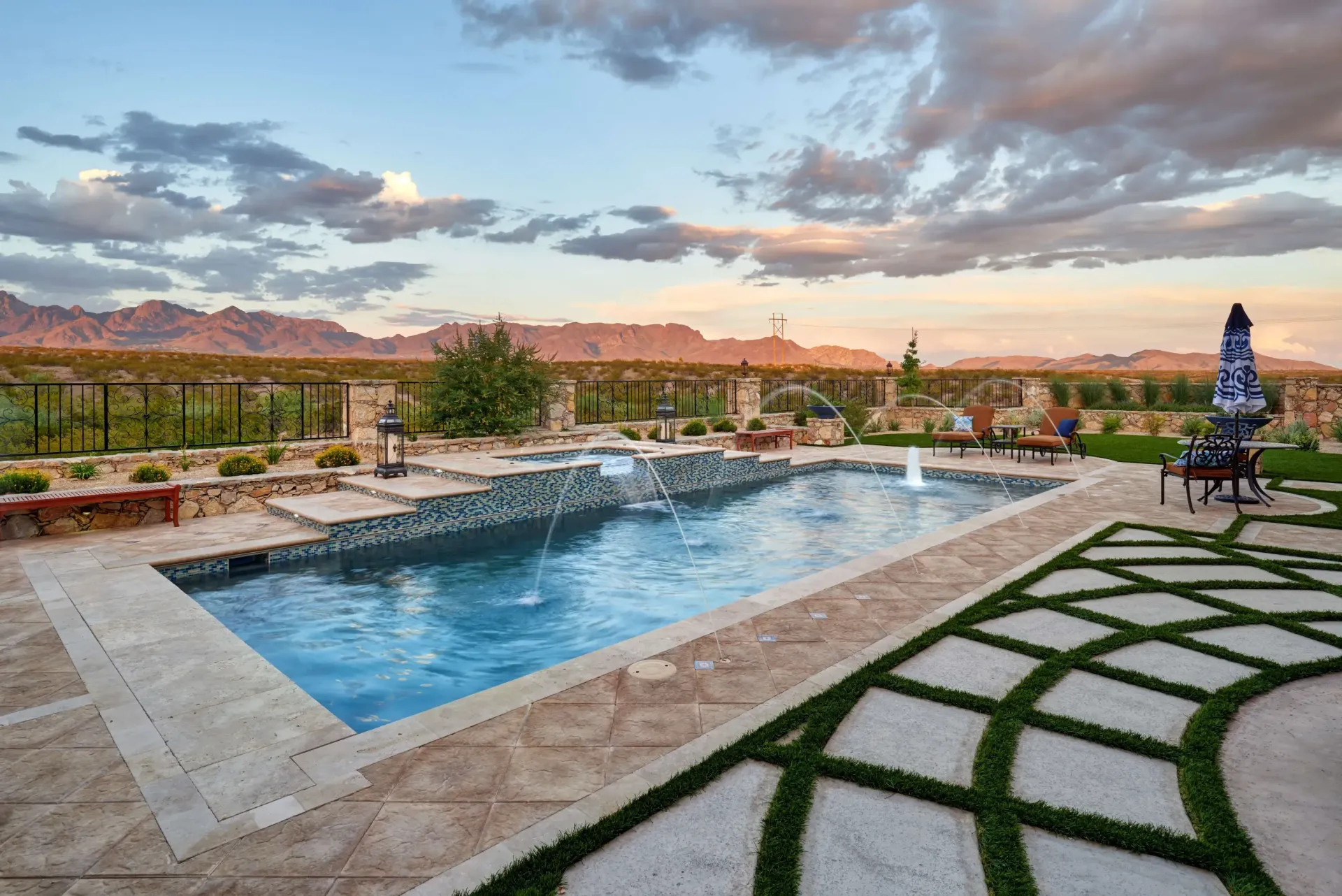 Swimming pool with water fountains, surrounded by landscaping, under a cloudy sunset sky.