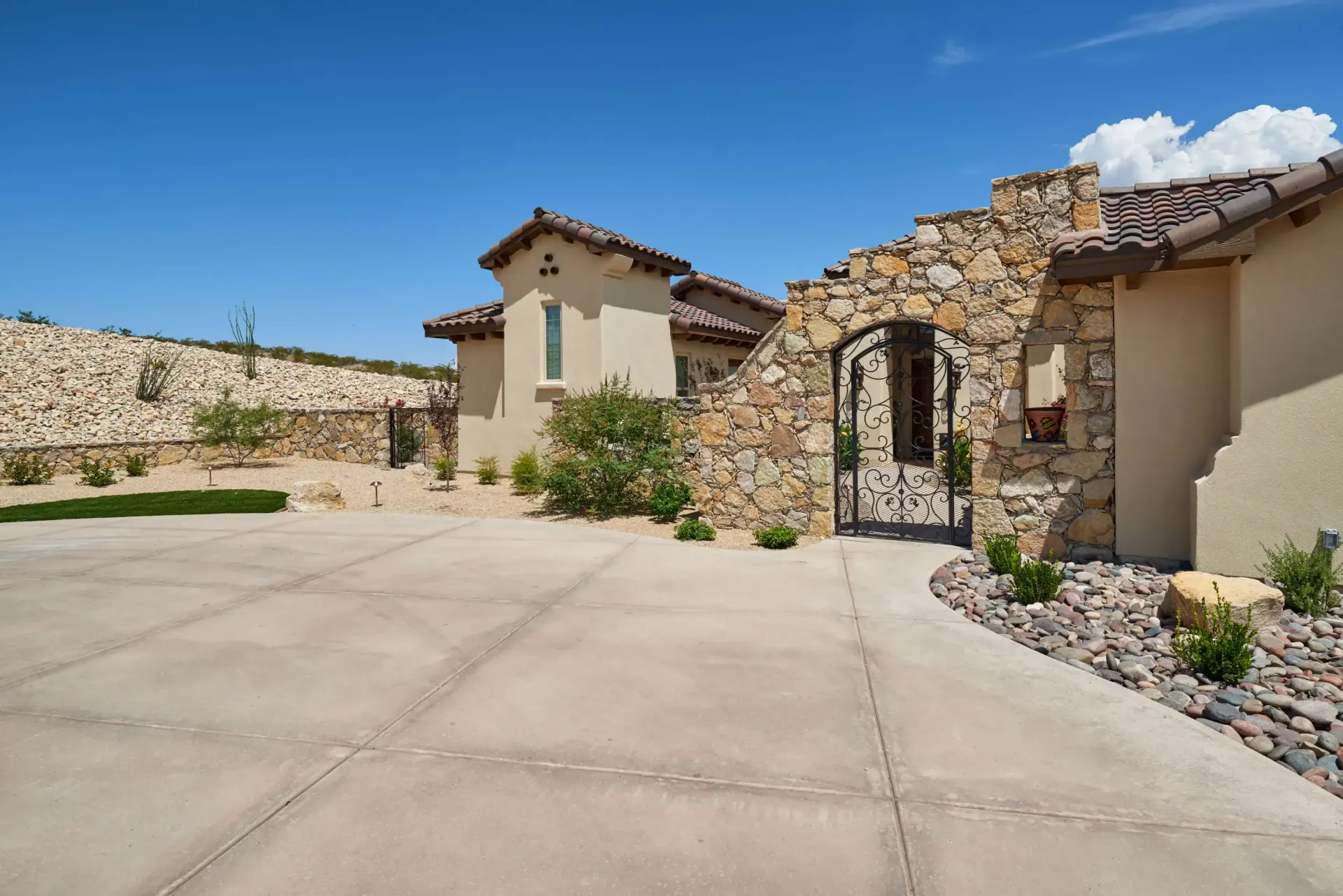 Stone archway leads to a house with beige walls and a tiled roof, under a blue sky.