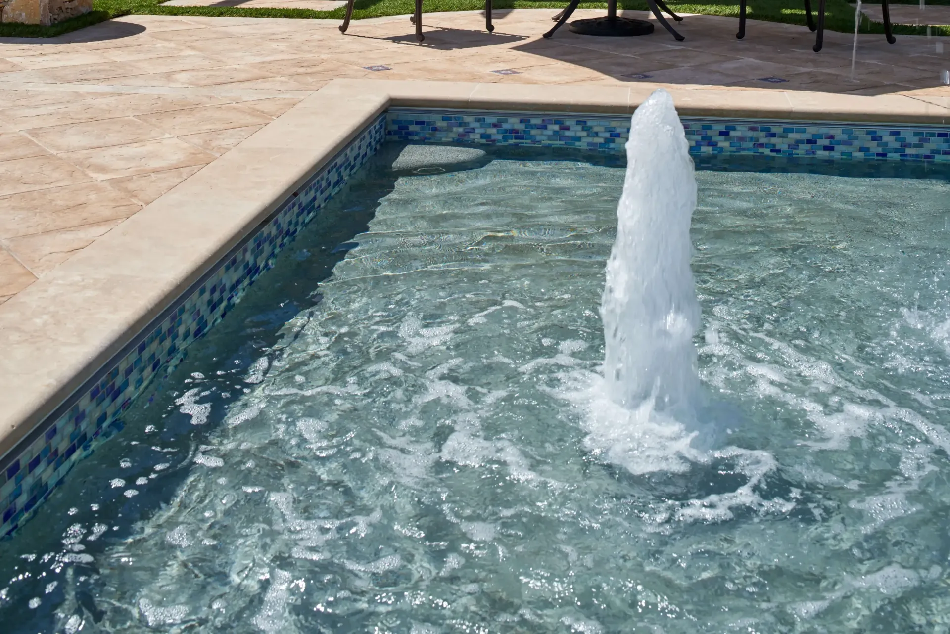 Fountain of water spouting in a small pool with blue mosaic tiles. Beige stone border, outdoor setting.