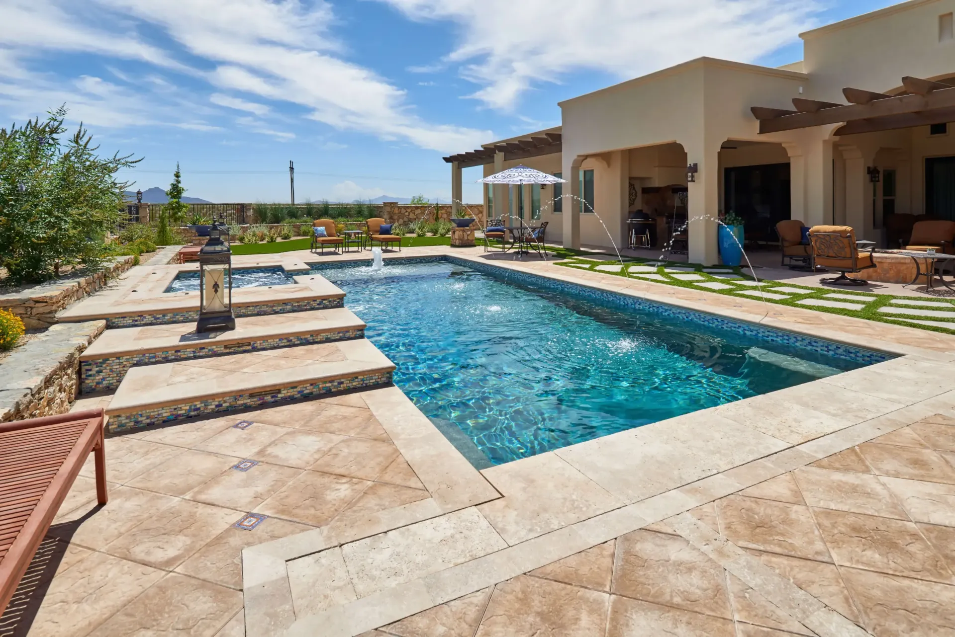Swimming pool with water fountain and hot tub, surrounded by stone patio, lawn, and house with pergola.