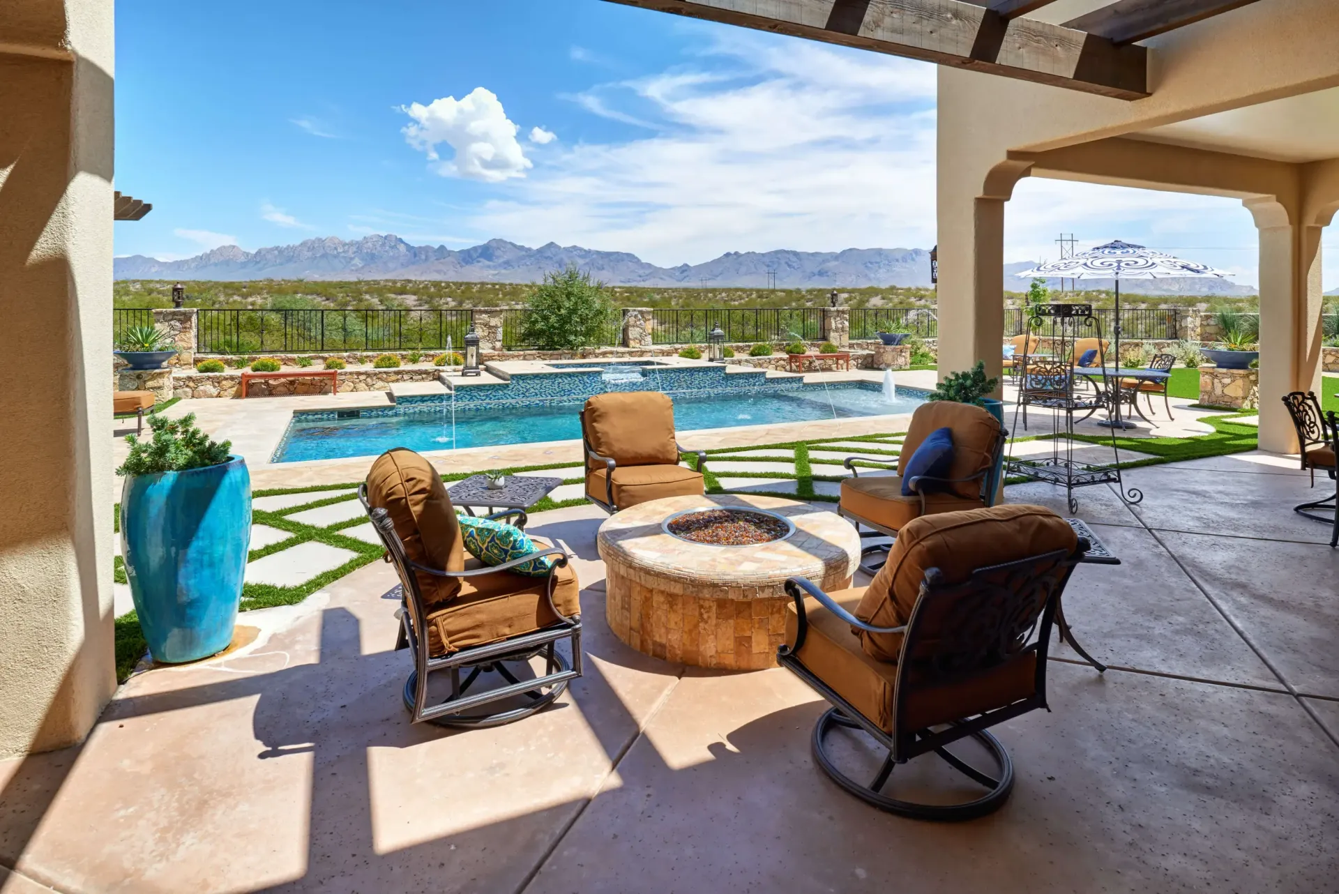 Patio with fire pit and seating, overlooking a pool and mountains under a blue sky.